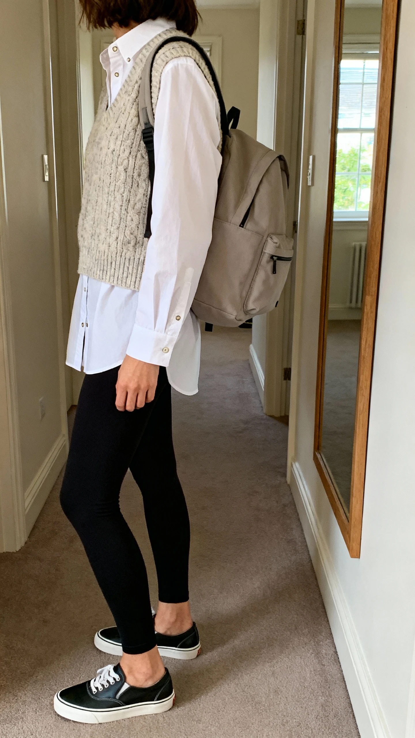 iPhone back/side view of a woman wearing a crisp white button-down layered under a knit vest, black leggings, sleek slip-on sneakers, and a minimalist backpack, face not visible, hallway mirror, natural window light, iPhone photo quality.