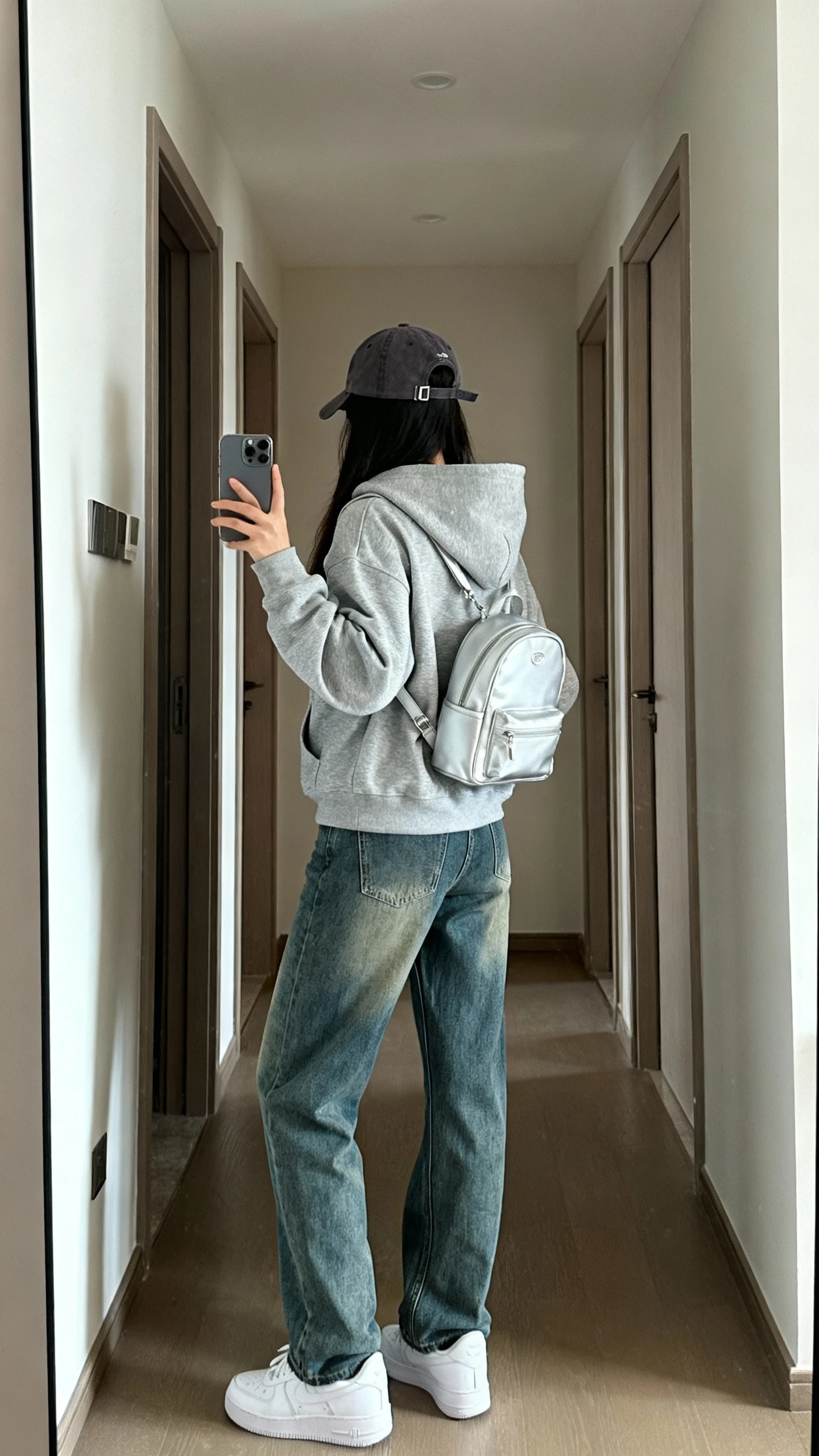iPhone back-view hallway mirror selfie of a woman wearing mom jeans, a heather gray hoodie, fresh white sneakers, baseball cap and small backpack, face not visible, natural daylight, casual iPhone photo