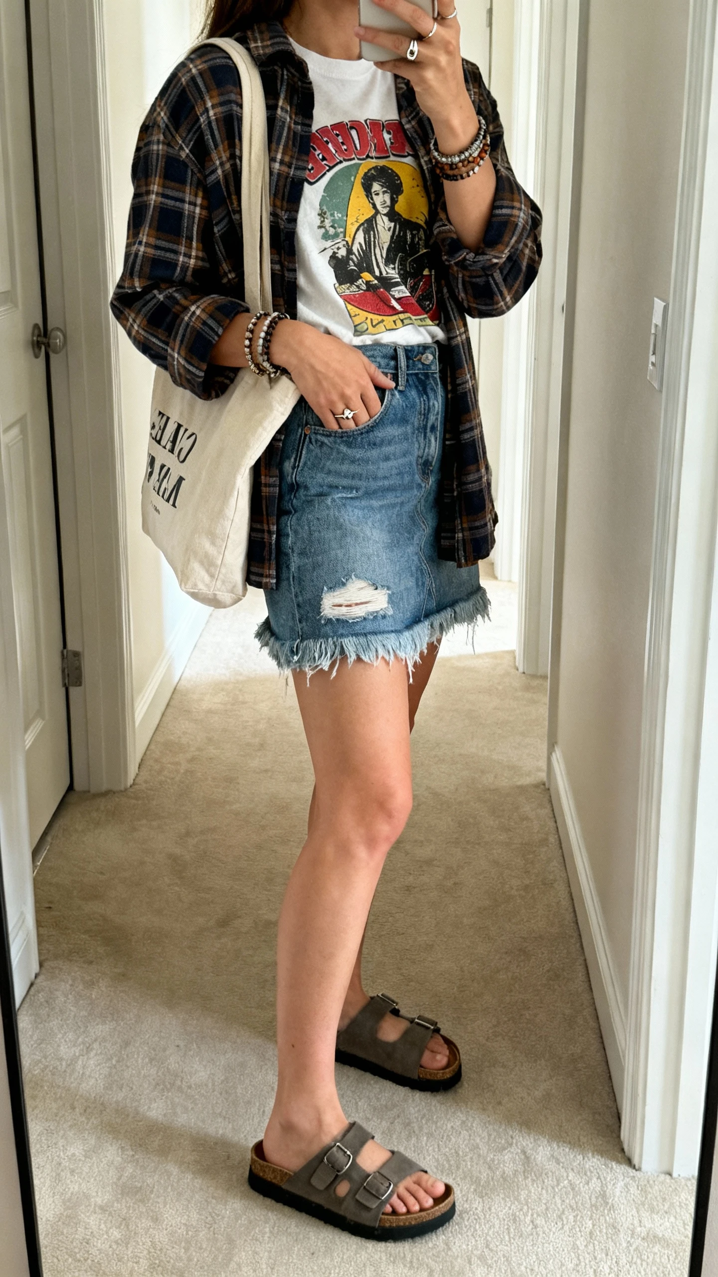 iPhone back-view hallway mirror selfie of a woman wearing a distressed raw-hem denim mini skirt, vintage-inspired graphic tee, plaid overshirt, chunky slide sandals, a canvas tote, layered bracelets, and a signet ring; face not visible, casual indoor daylight, iPhone photo quality.