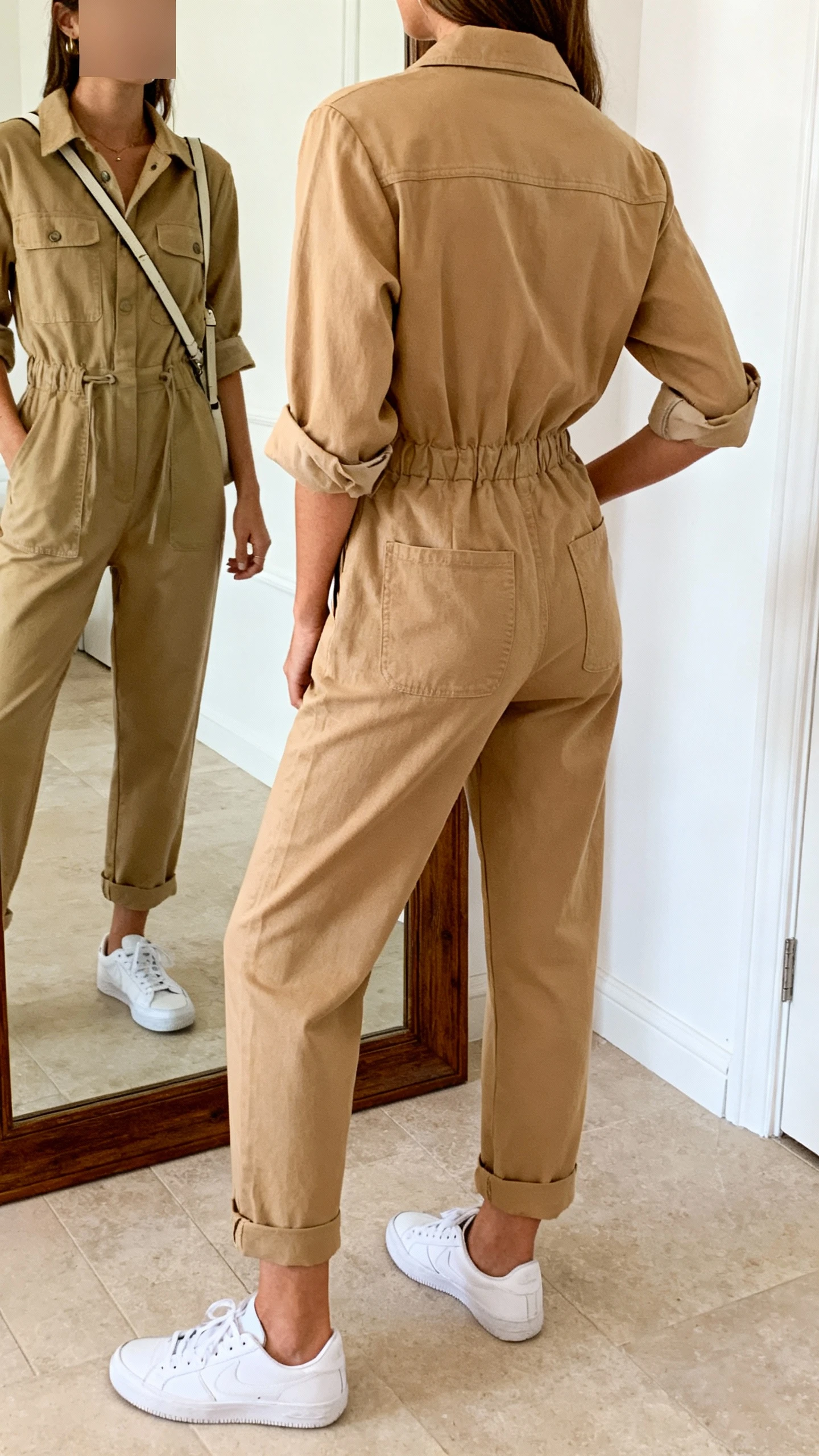 iPhone back-view mirror photo of a woman in a tan utility jumpsuit with rolled sleeves, white low-top sneakers, and a crossbody bag, face not visible, full-length mirror, soft indoor daylight, iPhone photo quality.