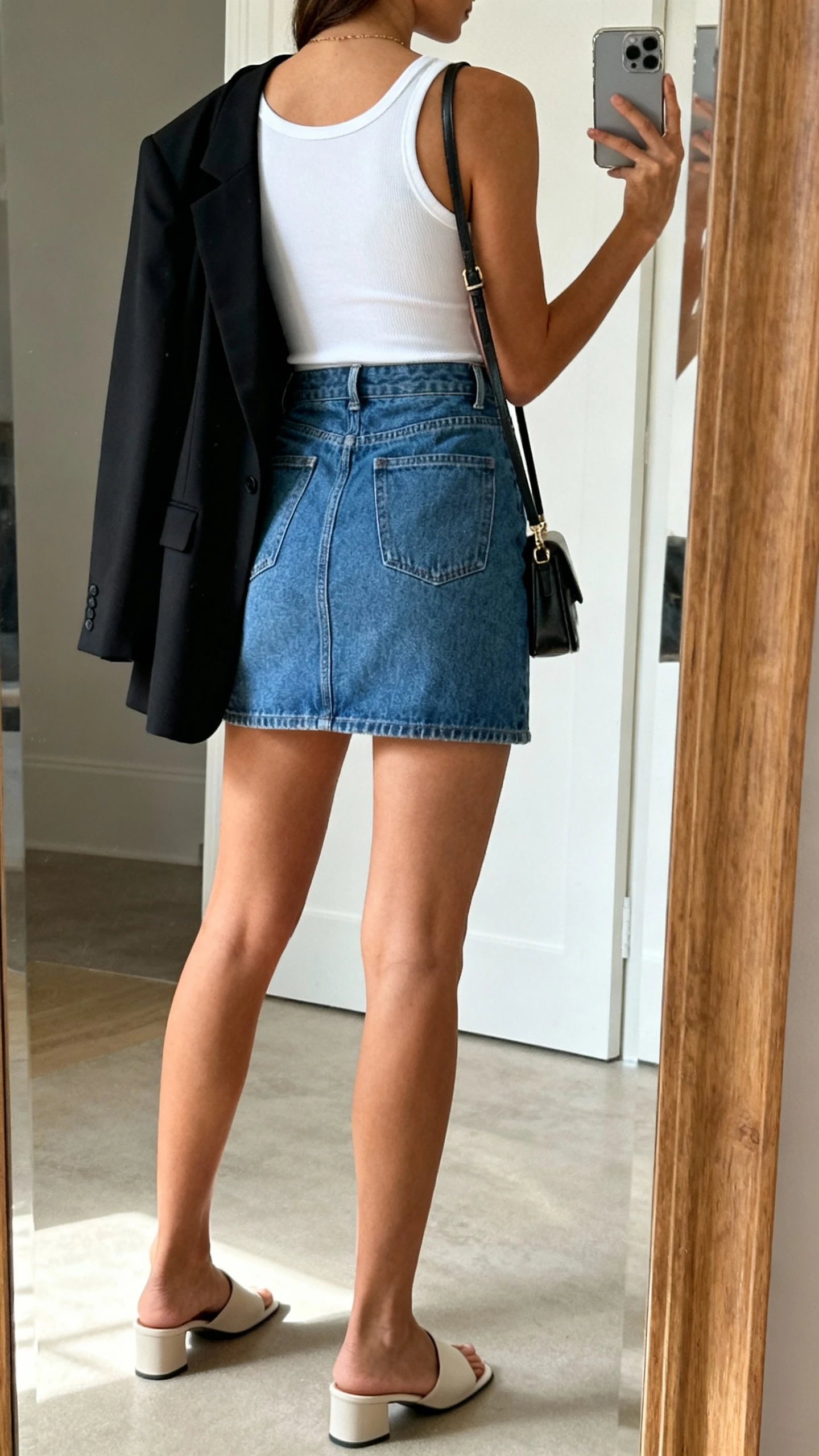 iPhone back-view mirror selfie of a woman in a fitted white tank, blue denim mini skirt, sleek black blazer draped on shoulders, and minimalist mules with a small shoulder bag, face not visible, full-length mirror, natural daylight, iPhone photo quality.