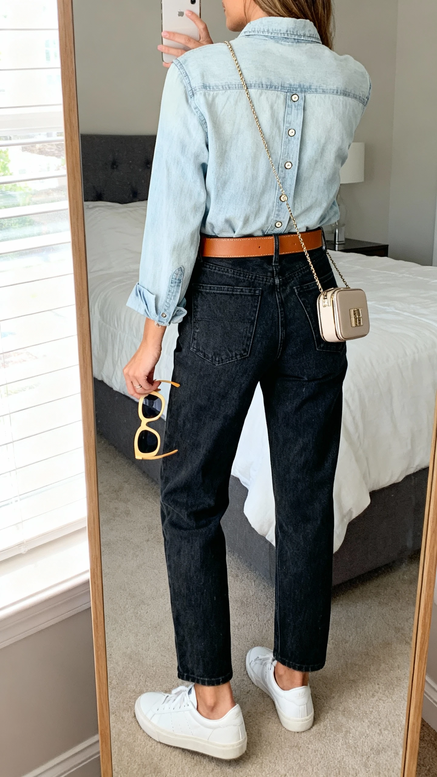 iPhone back-view mirror selfie of a woman in a light-wash denim button-up tucked into dark-wash mom jeans, tan leather belt, white sneakers, bold sunglasses in hand, and a compact crossbody, face not visible, bedroom mirror, bright window light, iPhone photo quality.