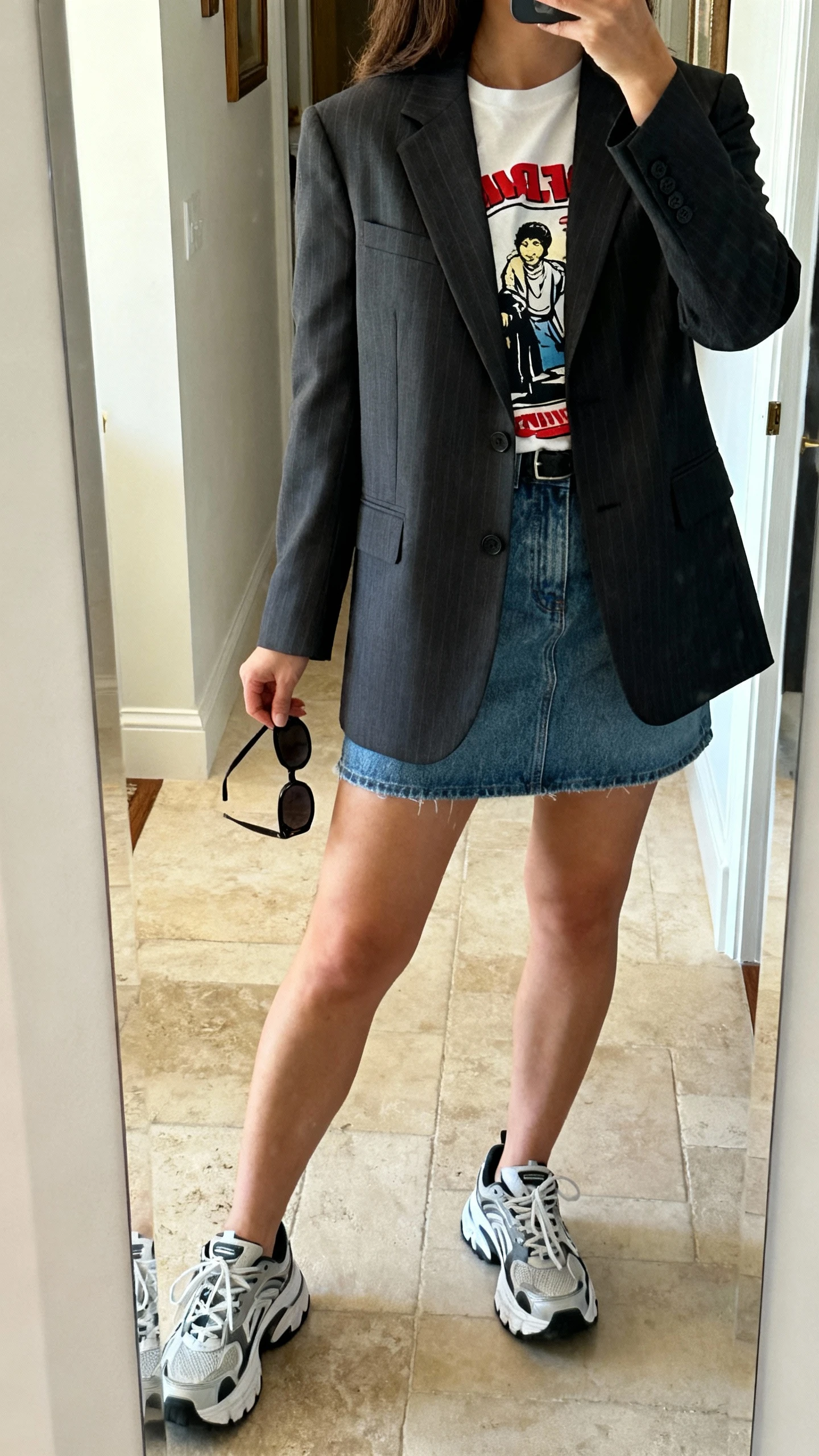 iPhone back-view mirror selfie of a woman in a tailored blazer over a graphic tee paired with a denim mini skirt, chunky sneakers, and sunglasses in hand, face not visible, entryway mirror, natural daylight, iPhone photo quality.