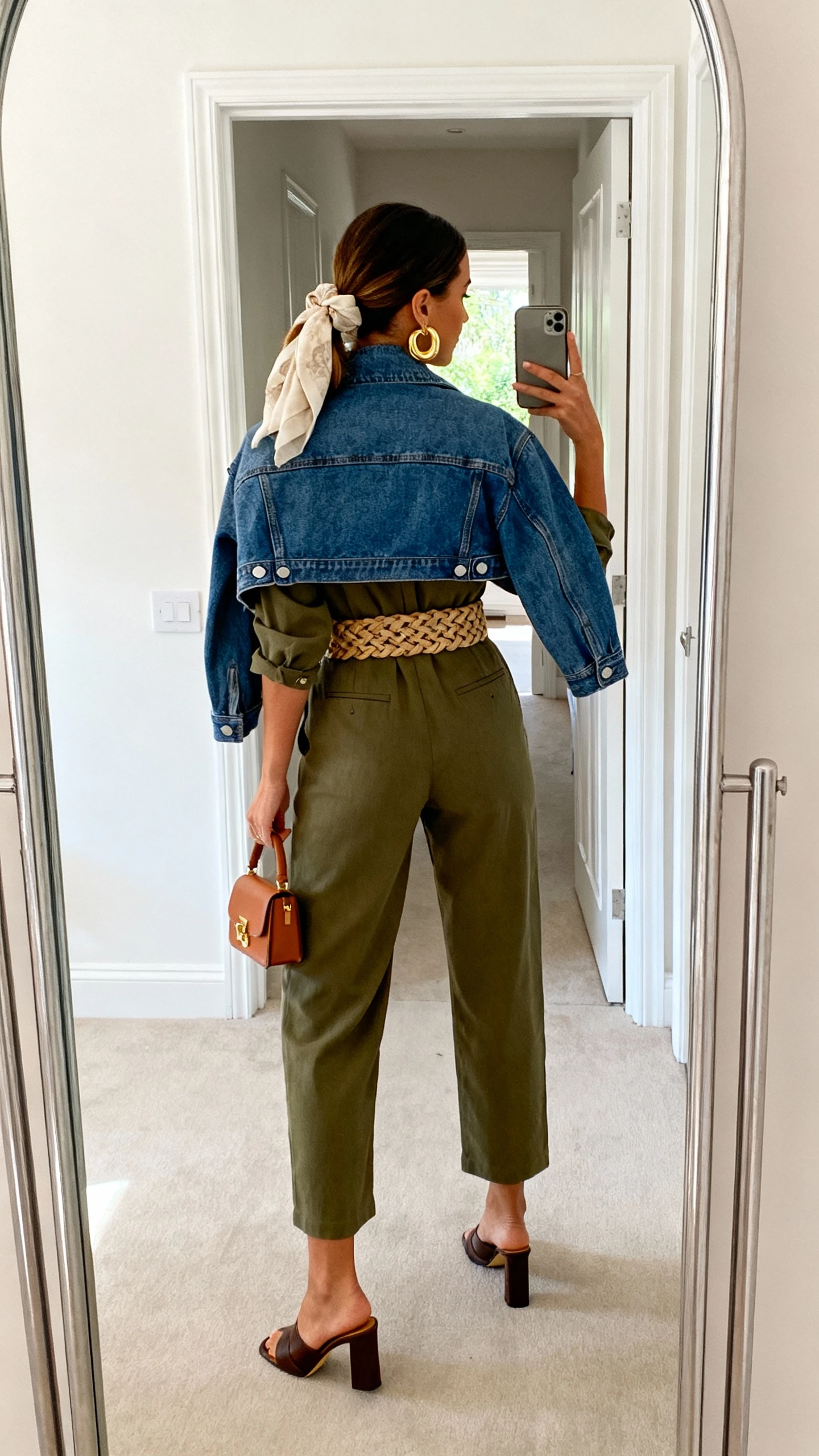 iPhone back view mirror selfie of a woman in an olive utility jumpsuit cinched with a woven statement belt, heeled mules, top-handle mini bag, chunky gold earrings, a light scarf tied in hair, and a cropped denim jacket draped over shoulders; face not visible, entryway mirror, natural daylight, iPhone photo quality.