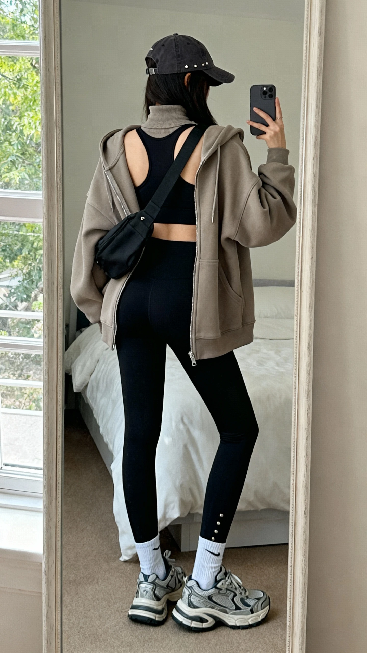 iPhone back view mirror selfie of a woman in matching black leggings and high-neck sports bra, oversized tonal zip hoodie, chunky dad sneakers, sling bag across chest, baseball cap, minimal studs, ankle socks visible; face not visible, bedroom mirror, natural window light, iPhone photo quality.