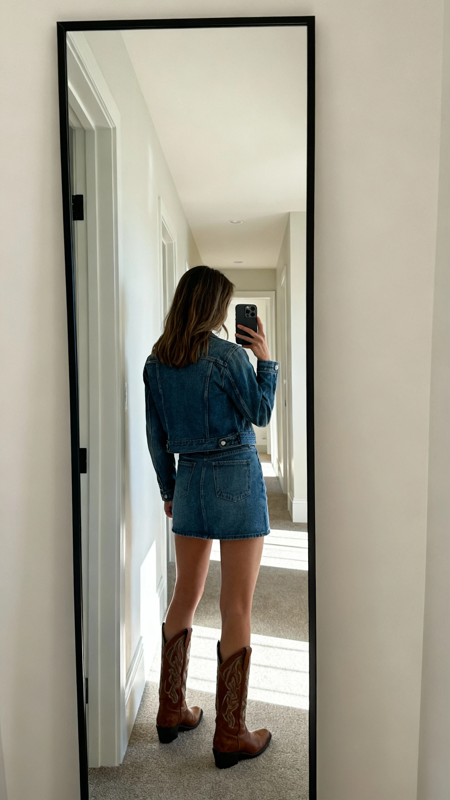 iPhone back-view mirror selfie of a woman wearing a denim jacket with a denim mini skirt and cowboy boots, face not visible, full-length mirror in hallway, natural daylight, iPhone photo quality.