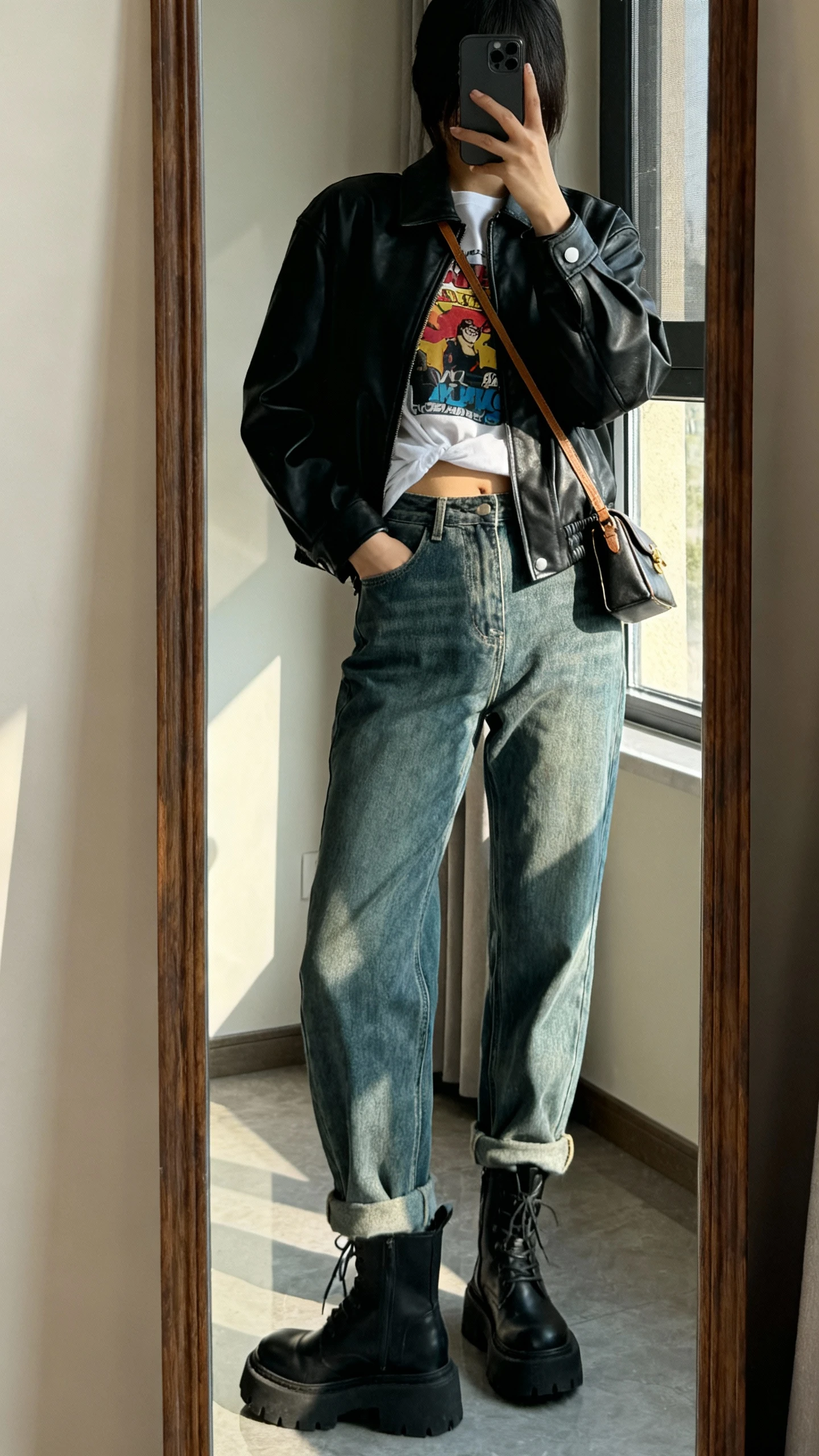 iPhone back-view mirror selfie of a woman wearing mom jeans, a graphic tee partially tucked, a black leather jacket, chunky boots, and a small shoulder bag, face not visible, full-length mirror, afternoon window light, iPhone photo quality.
