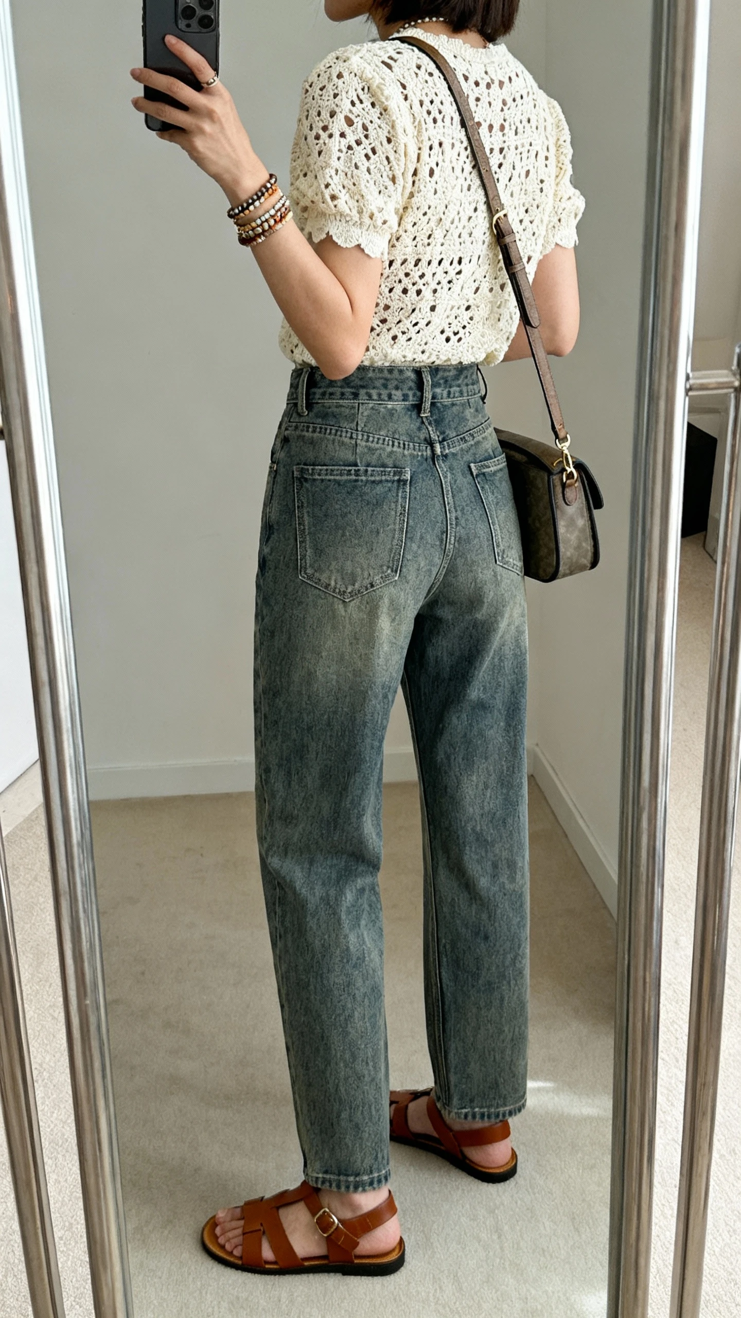 iPhone back-view mirror selfie of a woman wearing a cream crochet top with high-rise medium-wash denim, brown leather sandals, layered bracelets, and a structured crossbody, face not visible, full-length mirror, soft indoor light, iPhone photo quality.