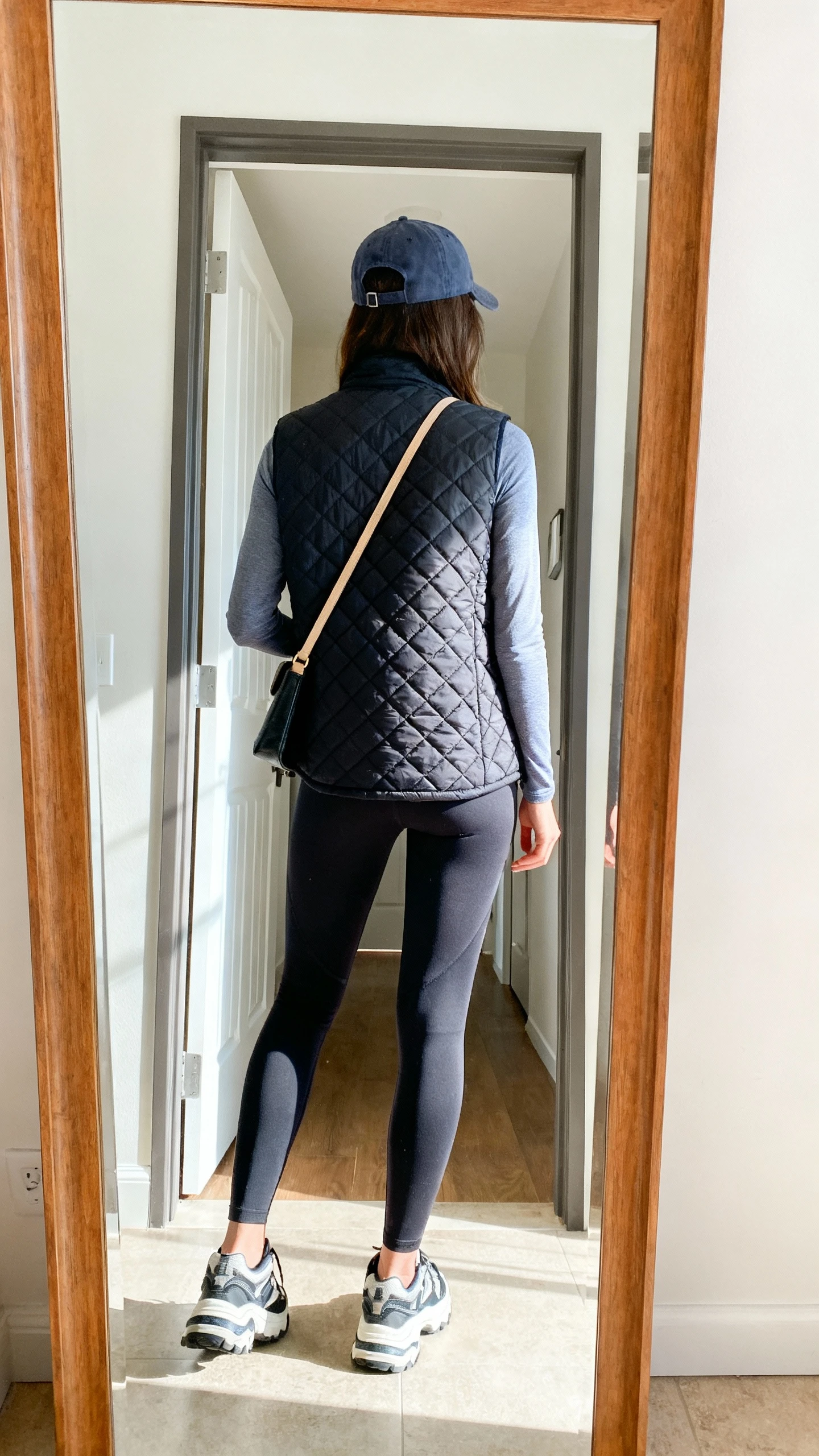 iPhone back-view mirror shot of a woman in a quilted vest layered over a long-sleeve base top and leggings, baseball cap, chunky sneakers, crossbody bag, face not visible, entryway mirror, bright daylight, iPhone photo quality.