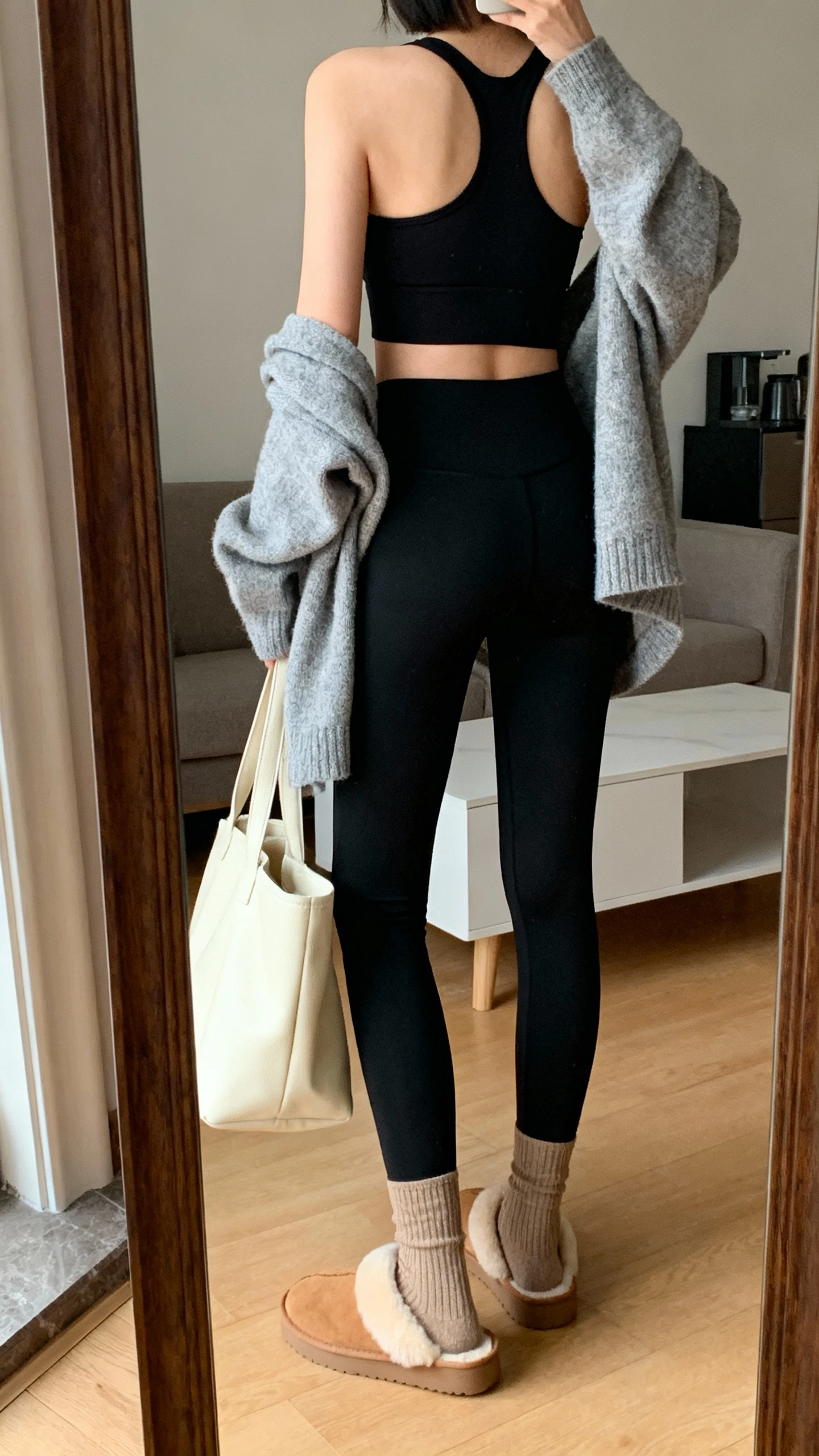iPhone back-view mirror shot of a woman in cozy athleisure: black leggings, black longline sports bra, soft heather-gray cardigan draped on top, tan shearling slides with socks, and a cream tote, face not visible, living-room mirror, warm indoor lighting, iPhone photo quality.
