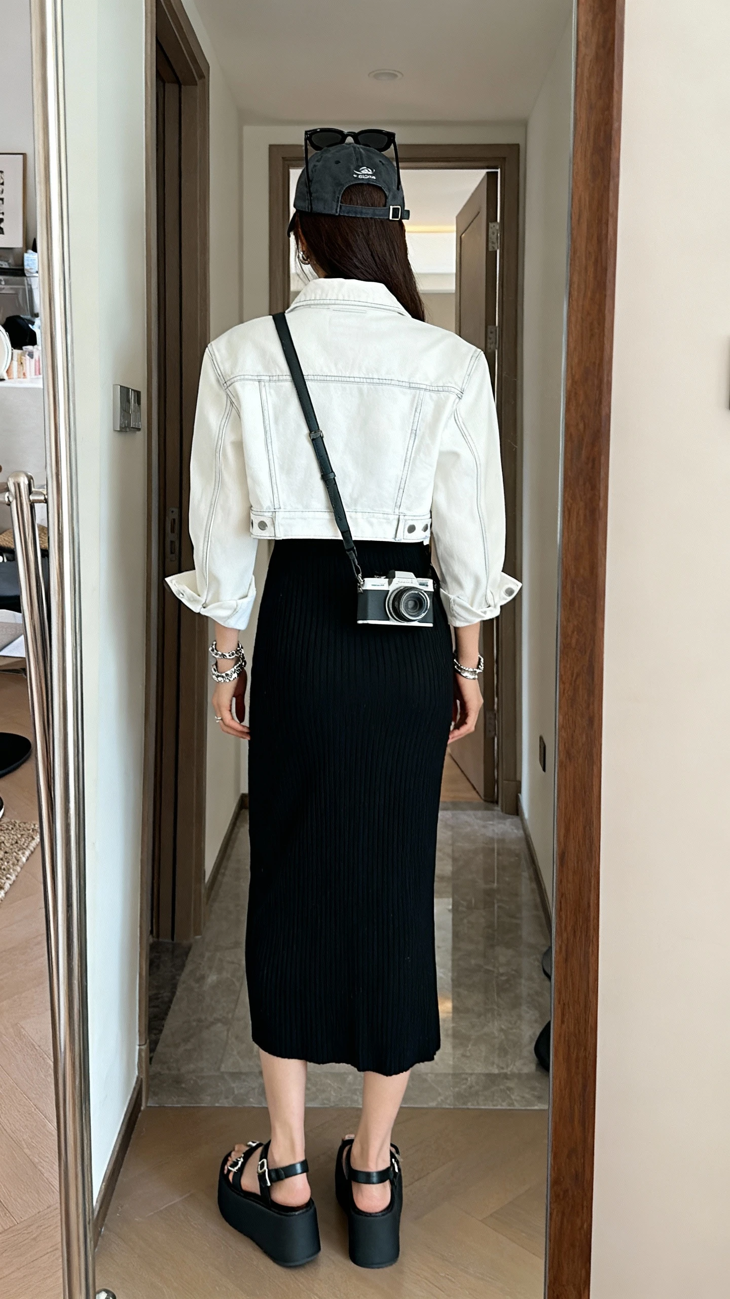 iPhone back-view mirror shot of a woman wearing a black ribbed midi dress with a cropped white denim jacket, platform sandals, a crossbody camera bag, black sunglasses perched on head and silver jewelry (optional baseball cap), face not visible, entryway mirror, indoor lighting, iPhone photo quality.
