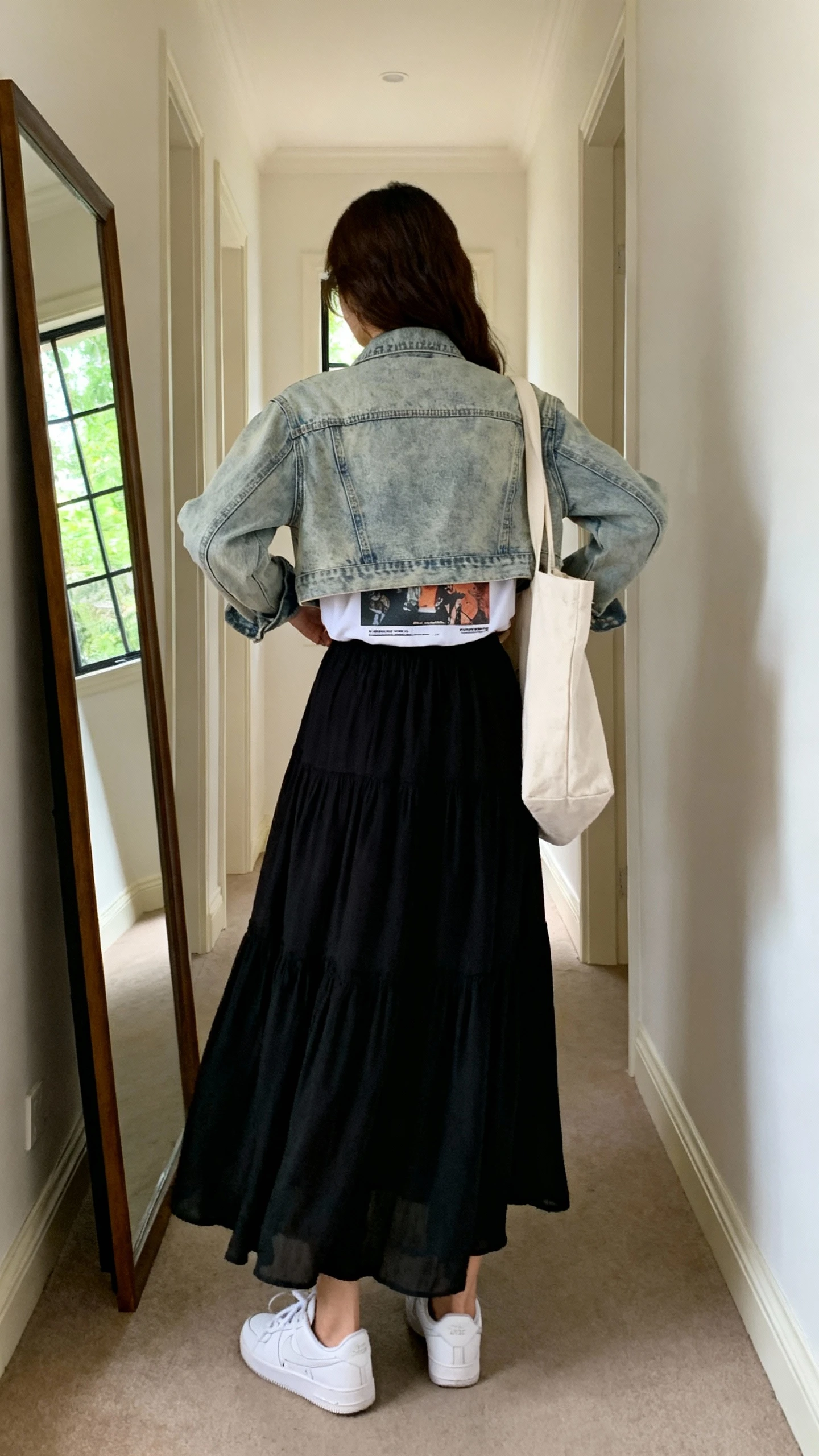 iPhone back view of a woman in a flowy black maxi skirt with a light-wash cropped denim jacket, graphic tee peeking underneath, white sneakers, canvas tote, face not visible, hallway full-length mirror, morning window light, iPhone photo quality.