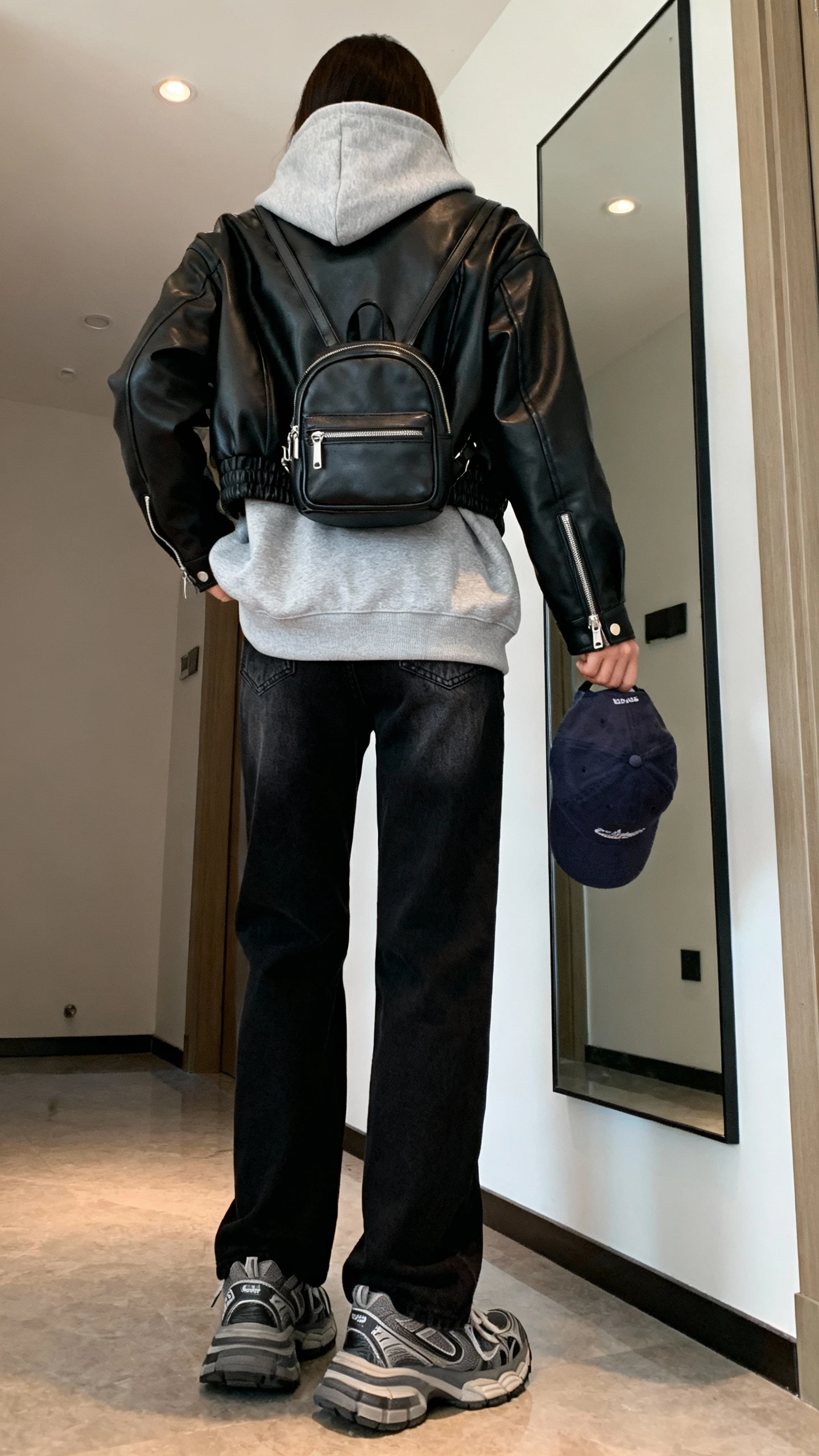 iPhone back view of a woman in a heather gray hoodie layered under a black leather moto jacket, straight-leg black jeans, chunky sneakers, mini backpack, baseball cap in hand, face not visible, hallway mirror, indoor lighting, iPhone photo quality.