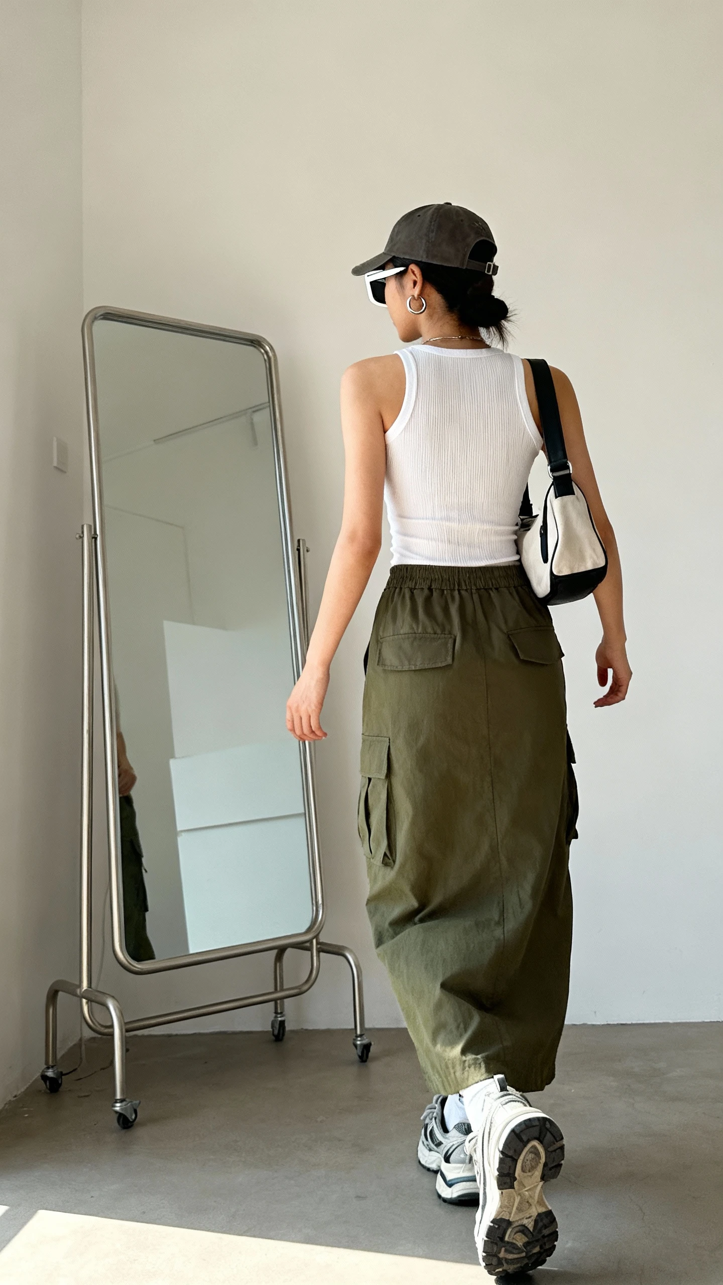 iPhone back view of a woman in an olive cargo maxi skirt, fitted white ribbed tank, chunky sneakers, sporty shoulder bag, rectangular sunglasses, huggie hoops, baseball cap; face not visible, walking pose toward full-length mirror, indoor daylight, casual iPhone photo quality.