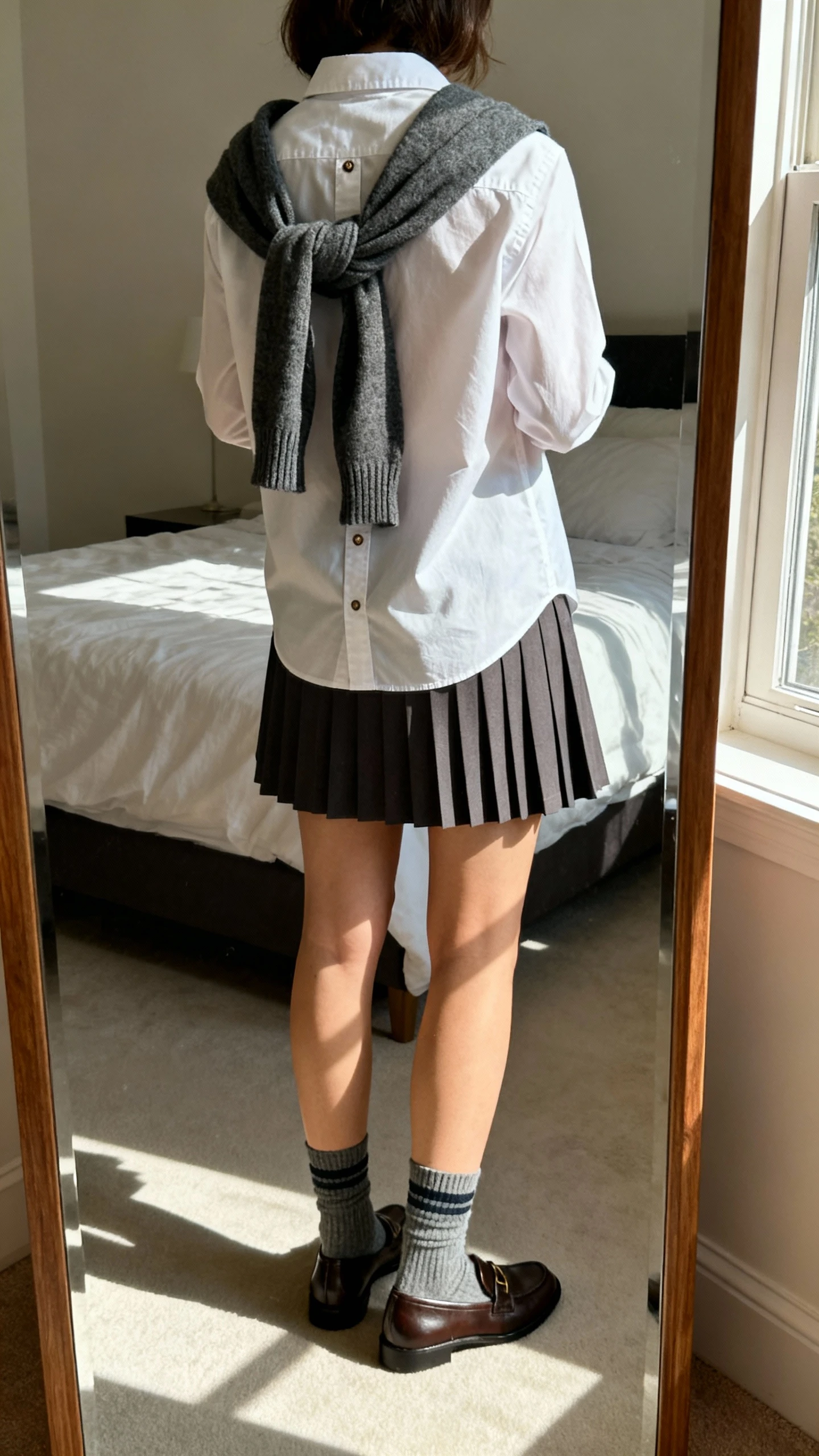 iPhone back view of a woman wearing a crisp button-down, a sweater tied over the shoulders, a pleated skirt, loafers, and crew socks, face not visible, full-length mirror in bedroom, natural window light, iPhone photo quality.