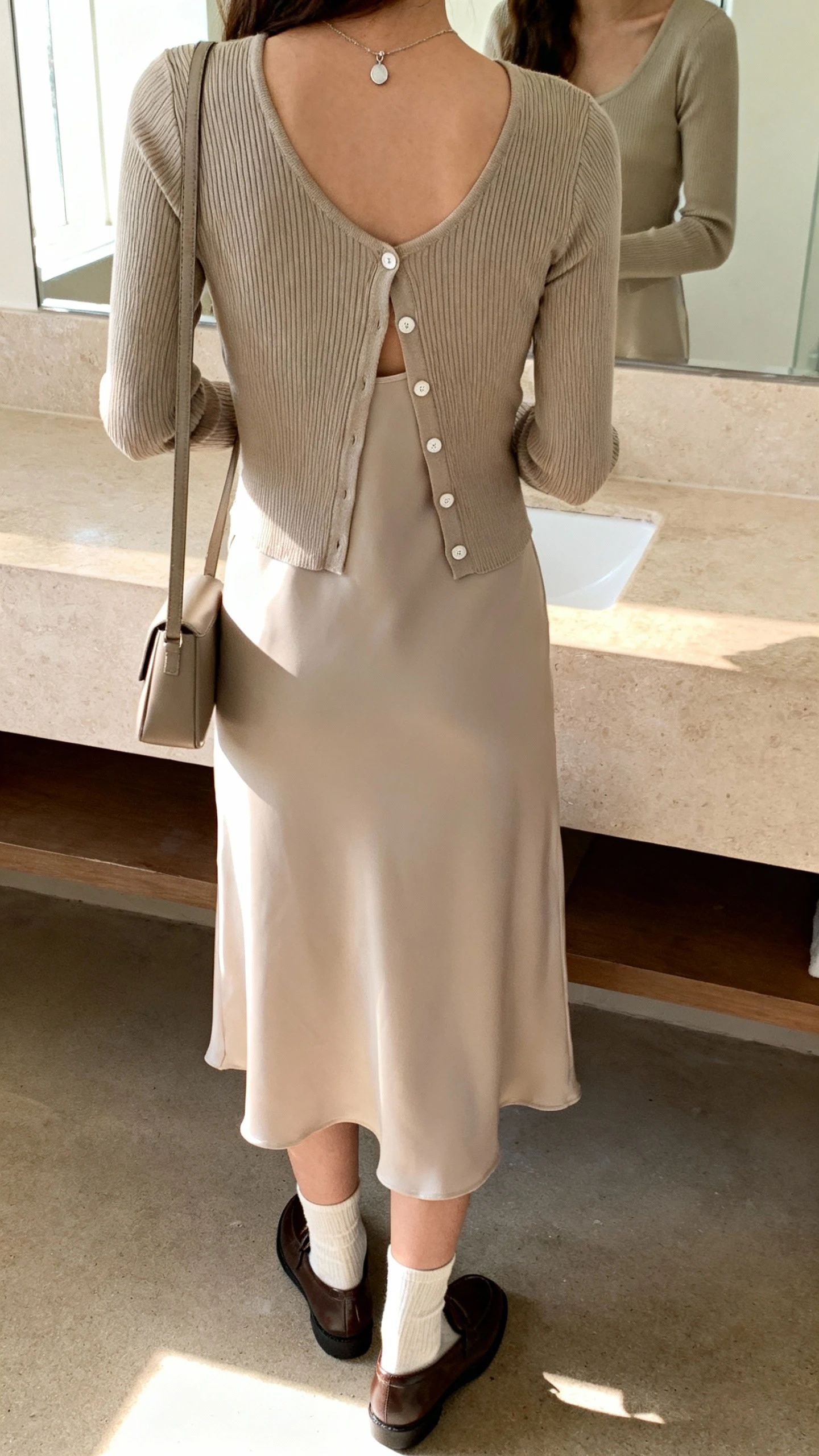 iPhone back view of a woman wearing a fitted cardigan buttoned halfway over a slip dress, ankle socks with loafers, slim shoulder bag, simple pendant necklace, face not visible, bathroom mirror, soft morning light, iPhone photo quality.
