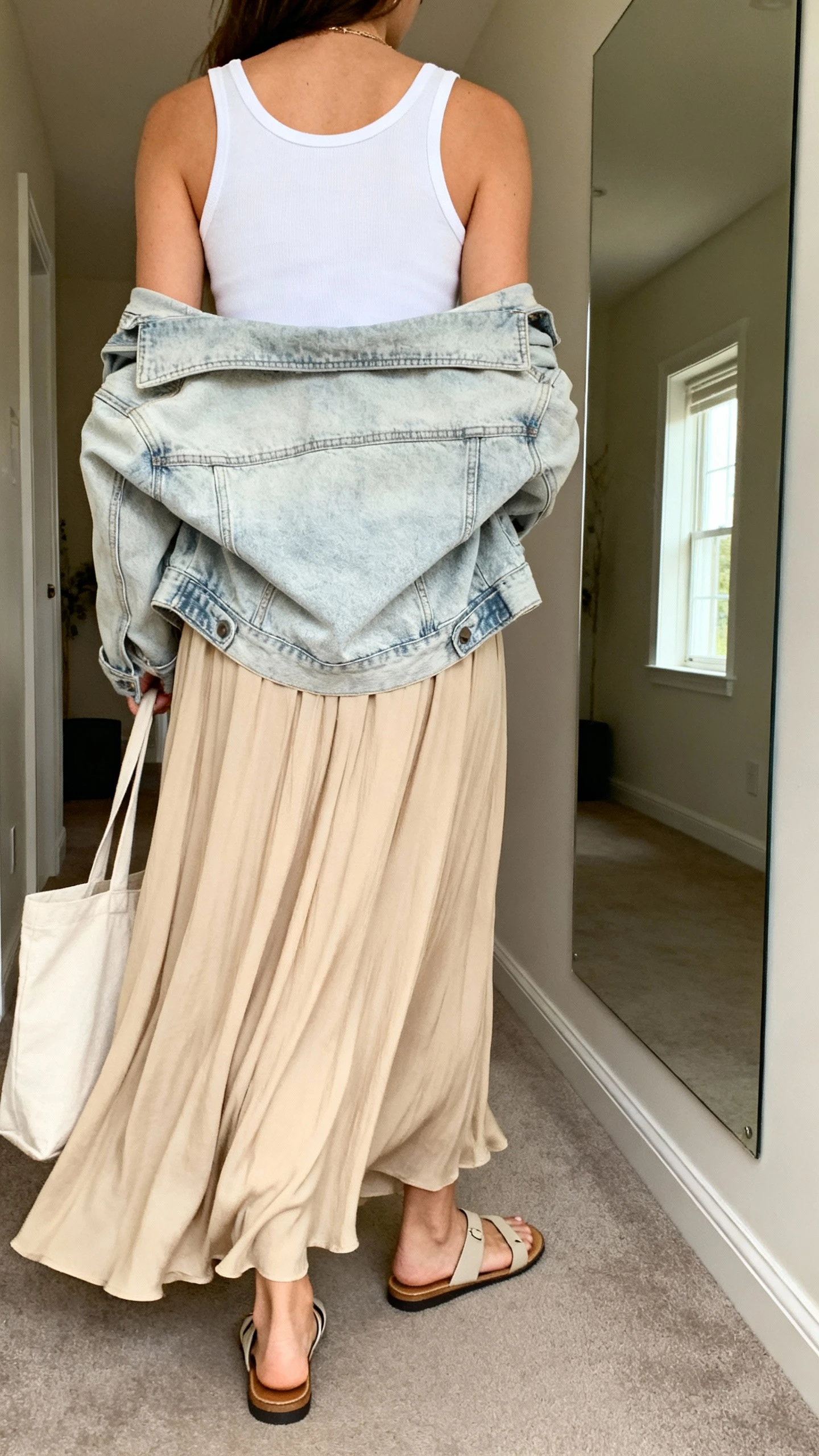 iPhone back view of a woman wearing a flowy beige maxi skirt with a white tank and a light-wash denim jacket draped over shoulders, flat sandals, tote bag, face not visible, hallway mirror, soft window light, iPhone photo quality