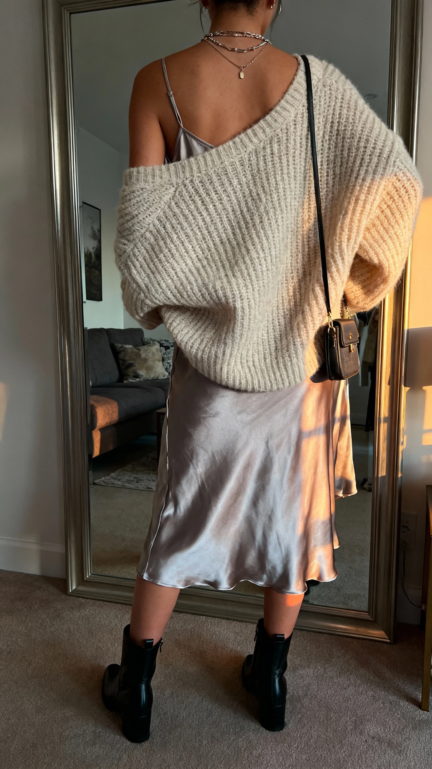 iPhone back view of a woman wearing a silky slip dress with an oversized chunky knit sweater on top, ankle boots, layered necklaces, small crossbody, face not visible, living room mirror, soft evening indoor light, iPhone photo quality.