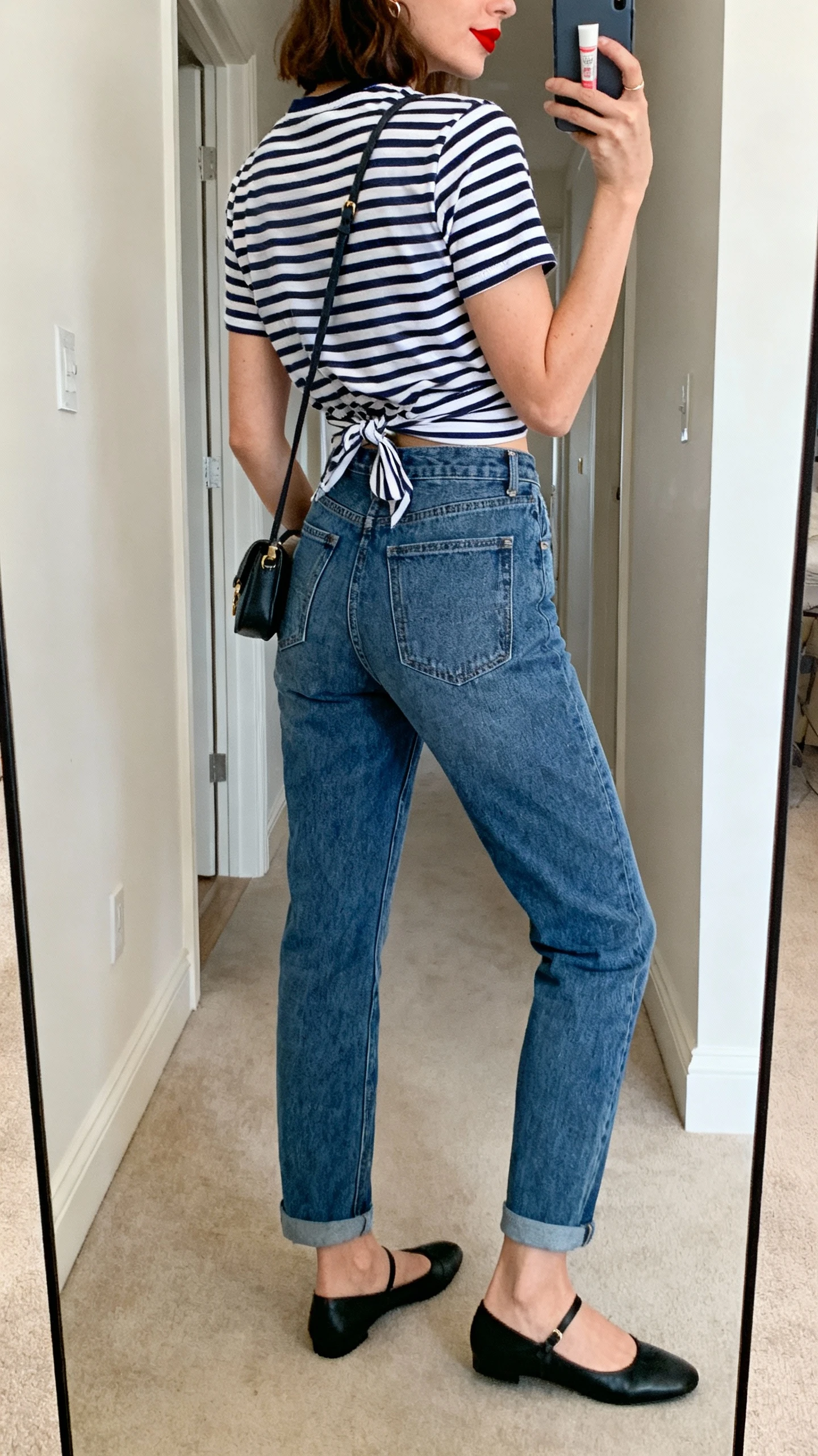 iPhone back view of a woman wearing mom jeans, classic navy-and-white striped tee half-tucked, black ballet flats, small crossbody bag, red lip seen only on hand-held tube (face not visible), hallway full-length mirror, natural daylight, iPhone photo quality