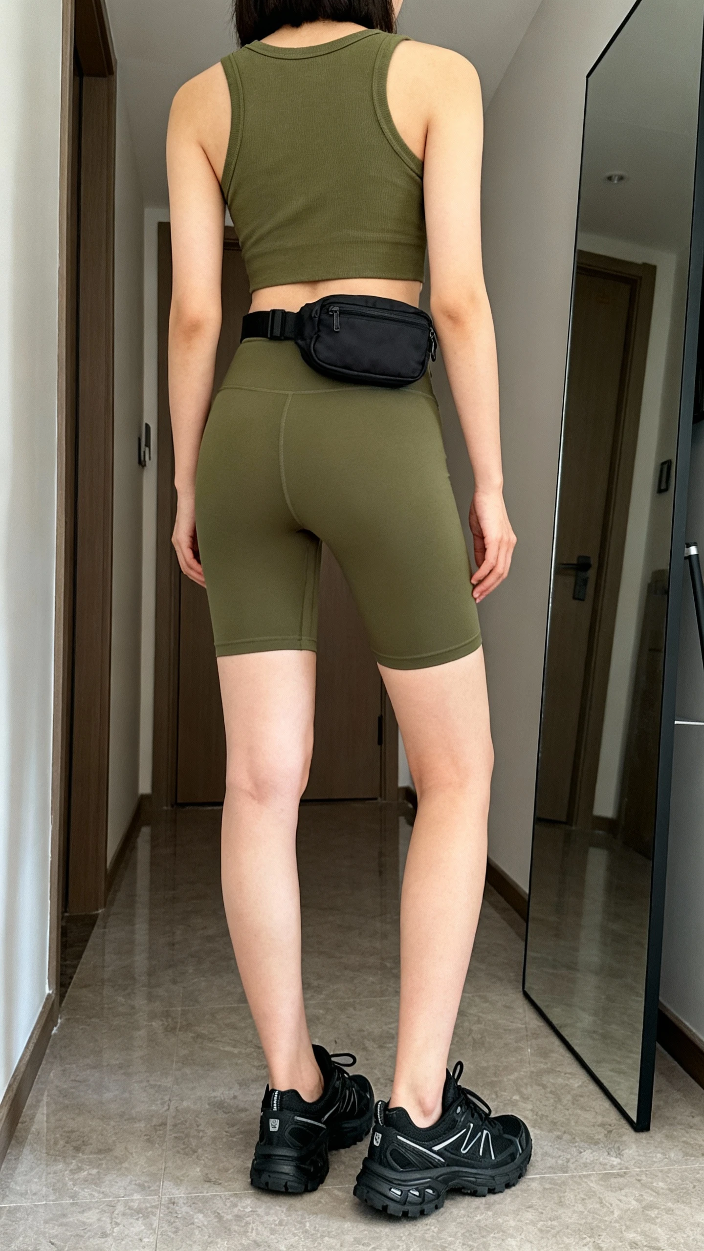 iPhone back view of a woman wearing olive green biker shorts, an olive green cropped tank, black cross-trainer sneakers, and a small black belt bag, face not visible, hallway mirror, natural daylight, iPhone photo quality.