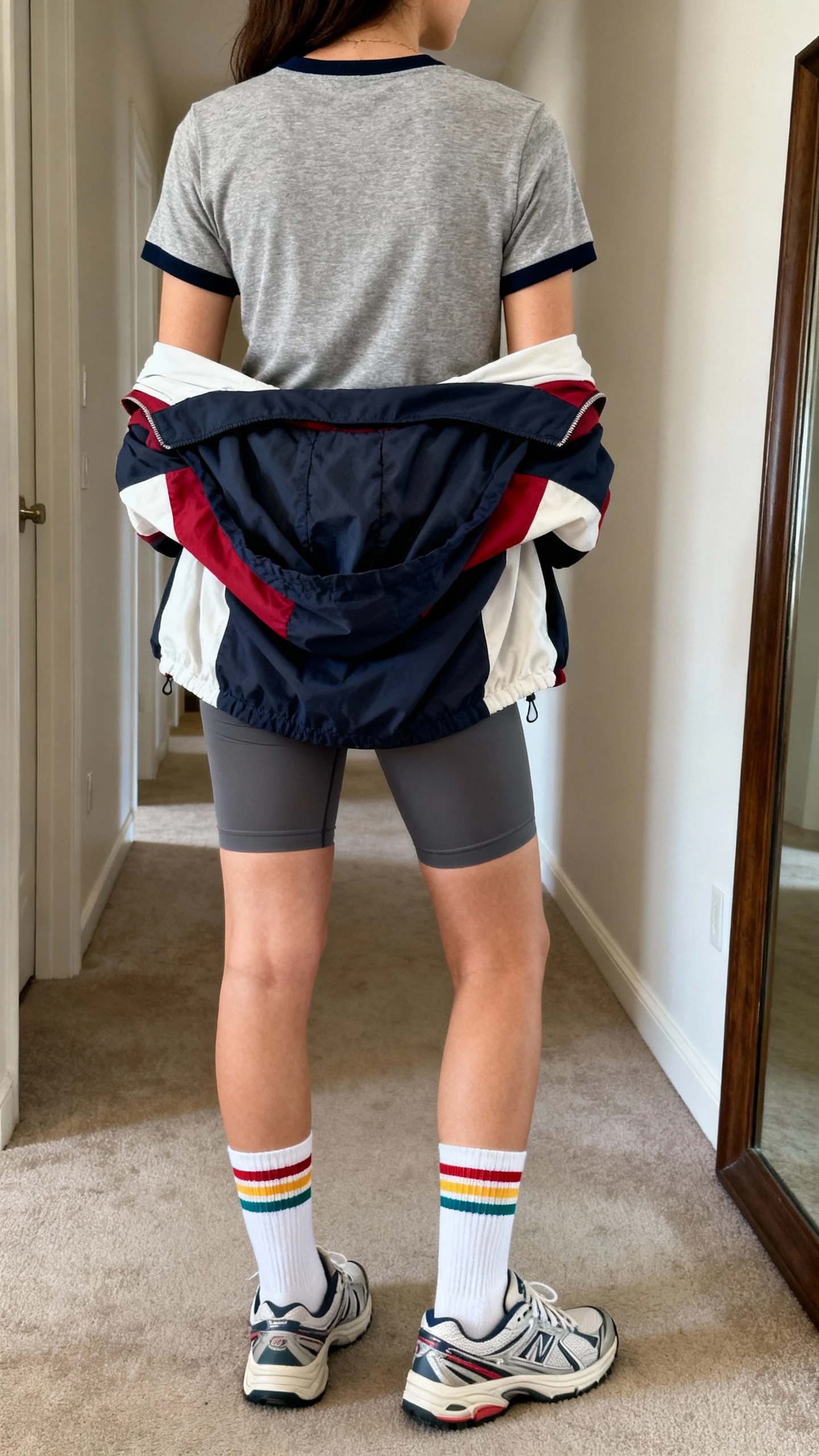 iPhone back view of a woman wearing retro runner vibes: heather gray ringer tee over a compressive bike short, white tube socks with colored stripes, vintage-style dad sneakers, and a color-block windbreaker draped over shoulders, face not visible, hallway full-length mirror, natural daylight, iPhone photo quality