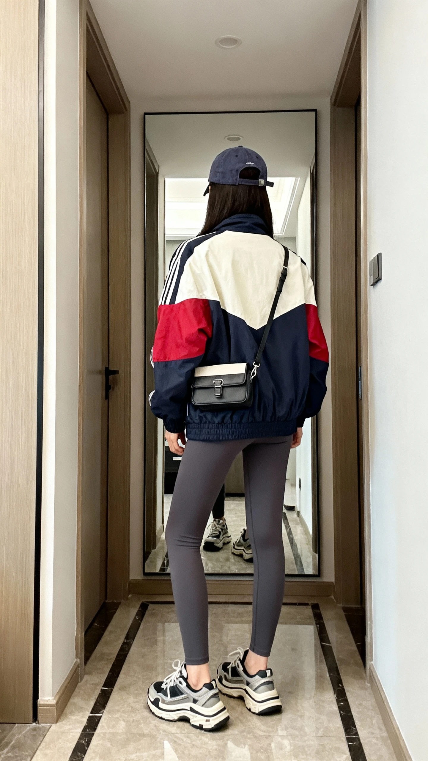 iPhone back view of a woman wearing sporty-chic outfit: statement bomber jacket (color-blocked), charcoal leggings, chunky dad sneakers, baseball cap, small belt bag worn crossbody, face not visible, hallway full-length mirror, indoor lighting, iPhone photo quality