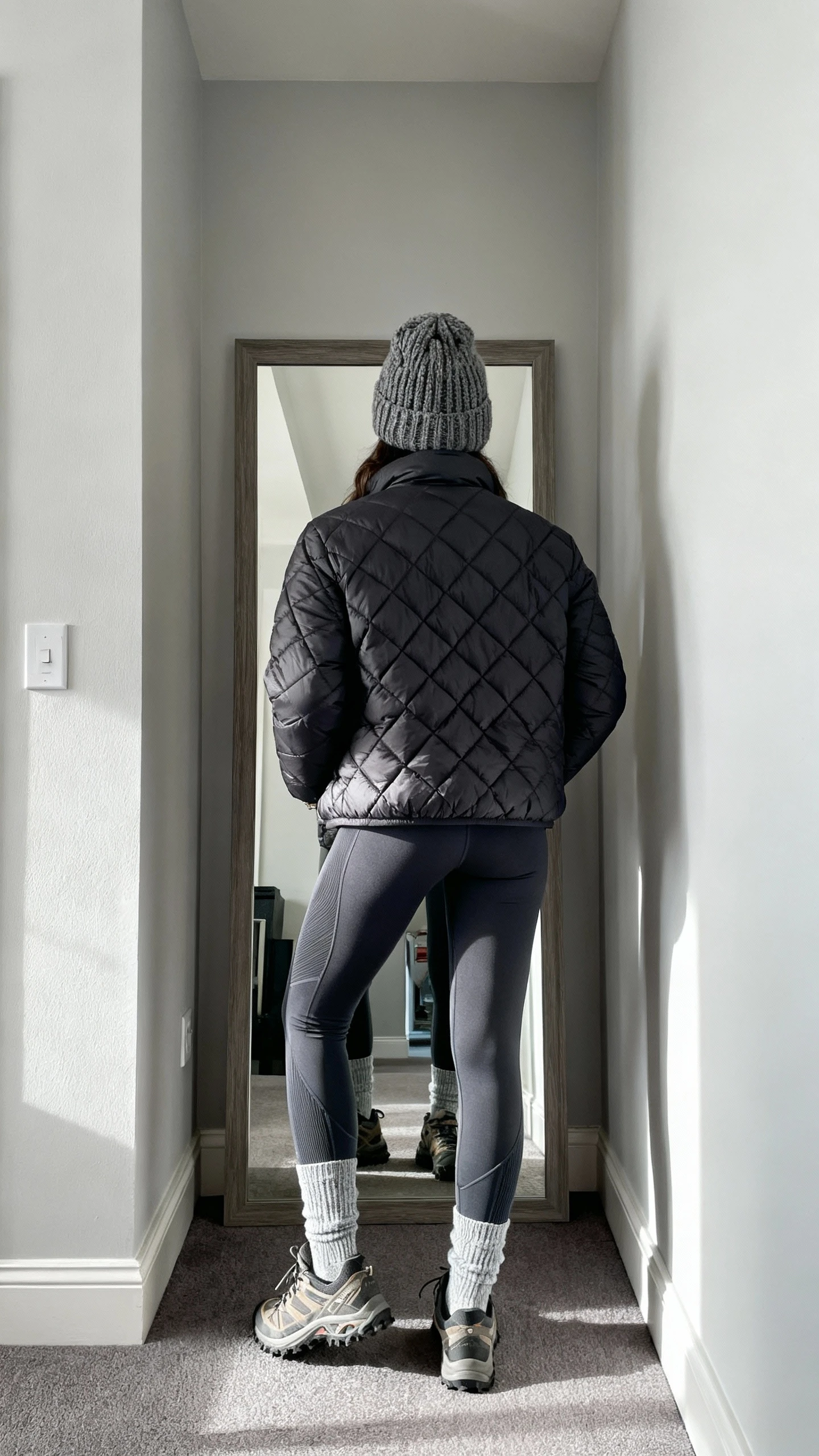 iPhone back view of a woman wearing thermal leggings, a quilted puffer jacket, chunky knit beanie, crew socks, and trail-style sneakers, face not visible, hallway full-length mirror, cool daylight from window, iPhone photo quality.