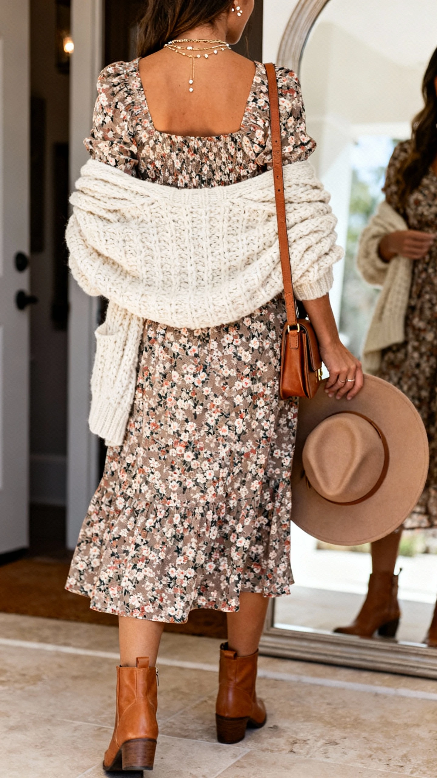 iPhone back-view photo of a woman wearing a floral midi day dress, chunky cream knit cardigan draped over shoulders, tan leather ankle boots, crossbody satchel, layered necklaces and delicate studs, with a wide-brim hat in hand, face not visible, entryway mirror, warm indoor light, iPhone photo quality.