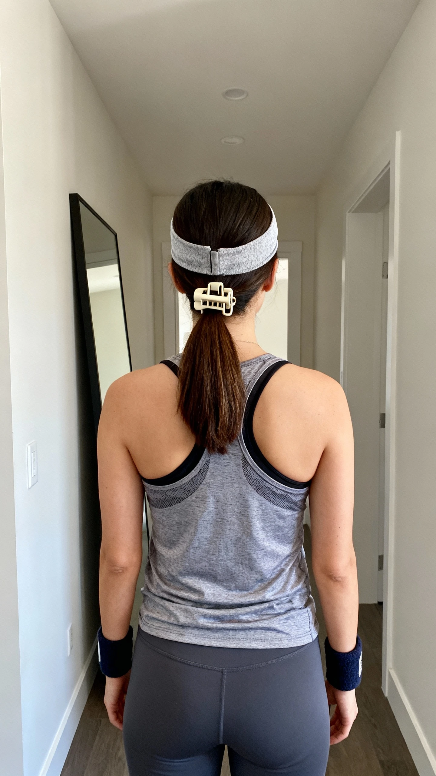 iPhone back-view photo of a woman wearing a no-slip headband, low ponytail with claw clip, wrist sweatbands, and a breathable tank with leggings, face not visible, full-length mirror in hallway, natural daylight, iPhone photo quality