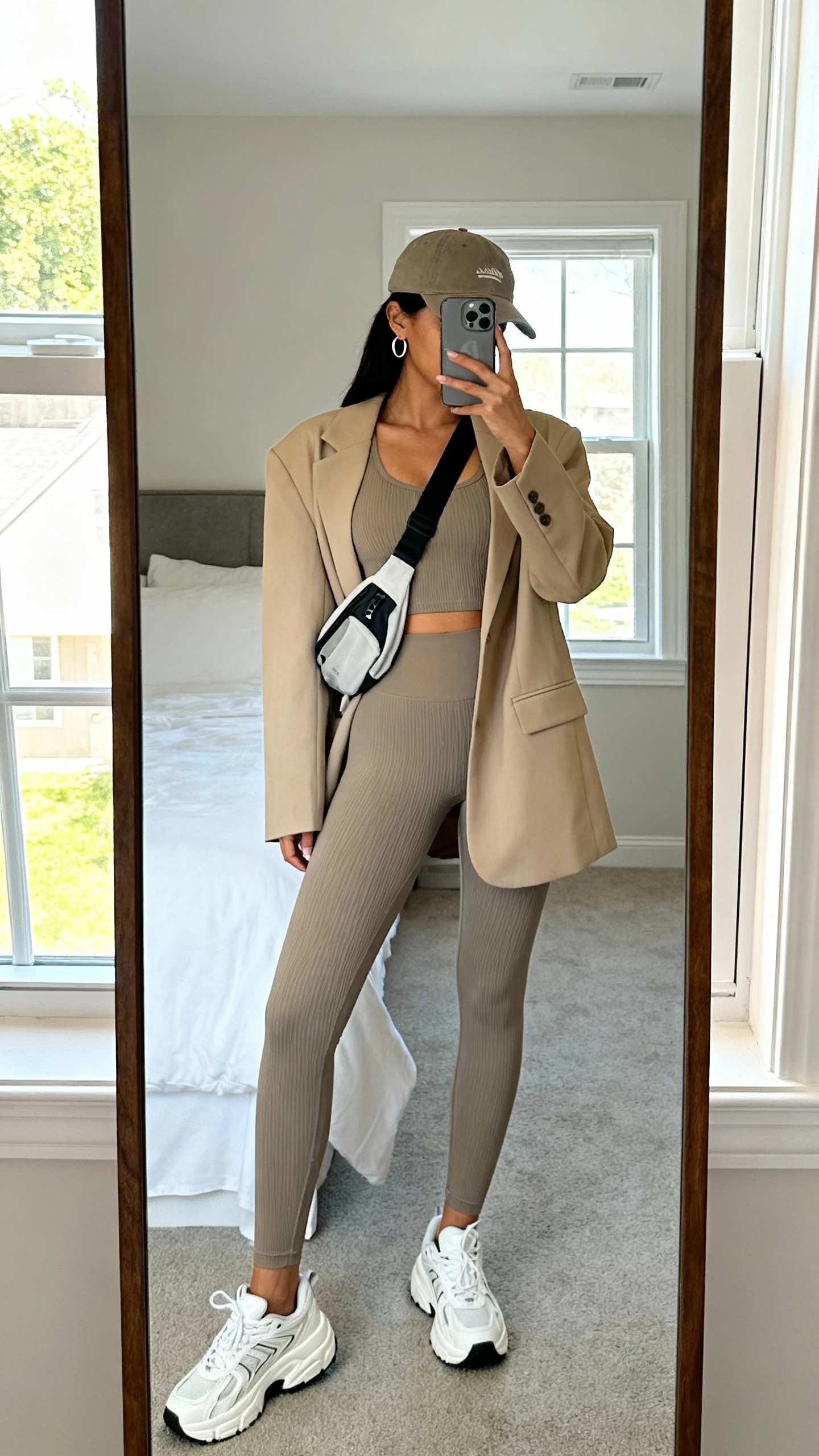 iPhone back-view selfie in a full-length mirror showing a woman in a matching ribbed tank and leggings, oversized beige blazer, chunky white sneakers, sporty crossbody, baseball cap, and minimal hoops; face not visible, bedroom mirror, bright window light, iPhone photo quality.