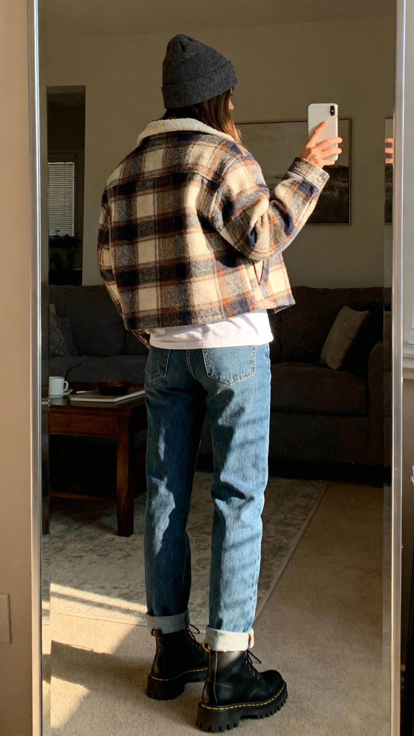 iPhone back-view selfie of a woman wearing a cozy plaid shacket over a tee with mom jeans, beanie, and lug-sole boots, face not visible, back turned to mirror, full-length mirror in living room, late afternoon natural light, iPhone photo quality.