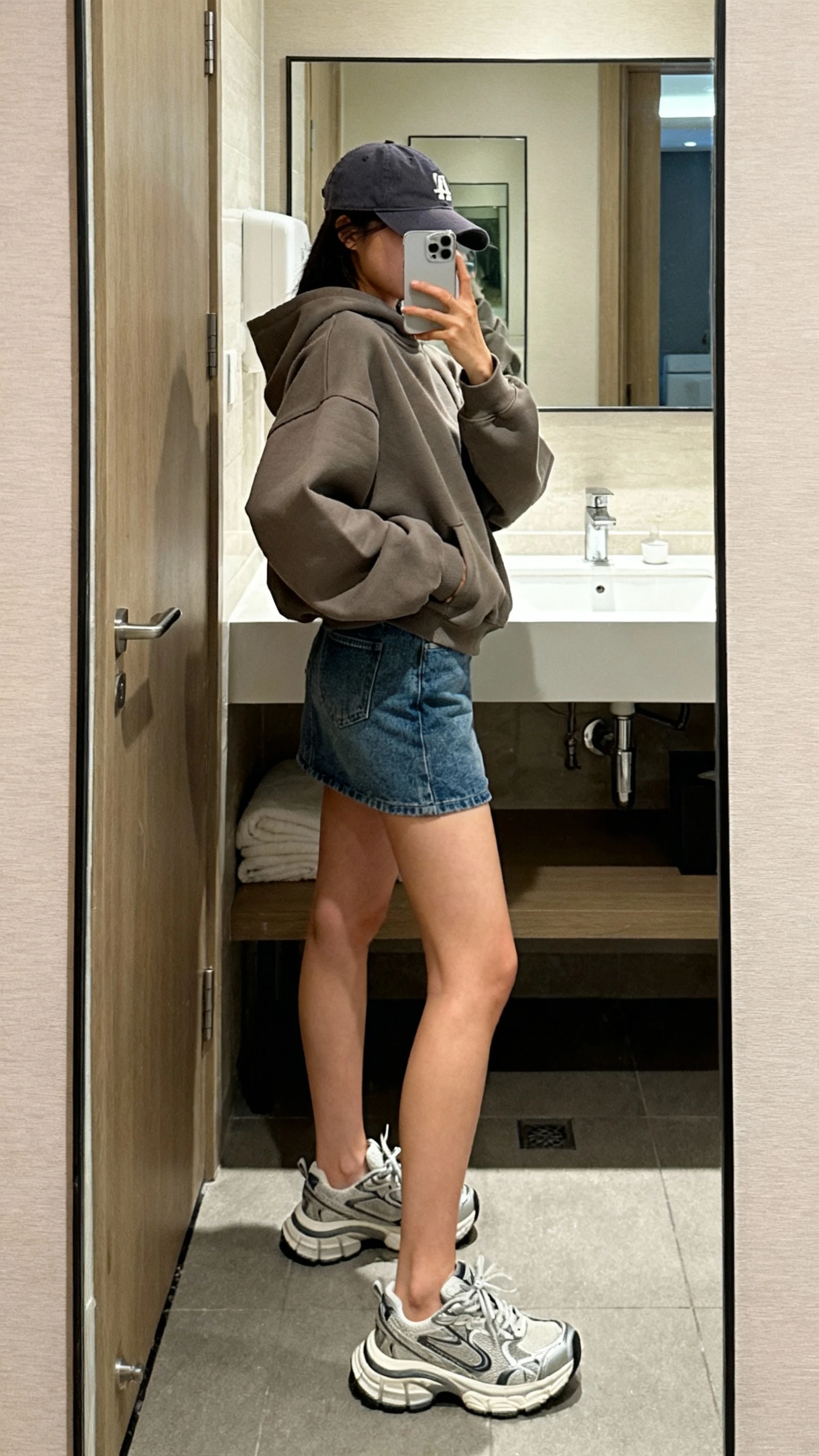 iPhone bathroom mirror selfie of a woman in a boxy hoodie with a denim mini skirt, chunky dad sneakers, and a baseball cap, face not visible, side angle, indoor lighting, iPhone photo quality.