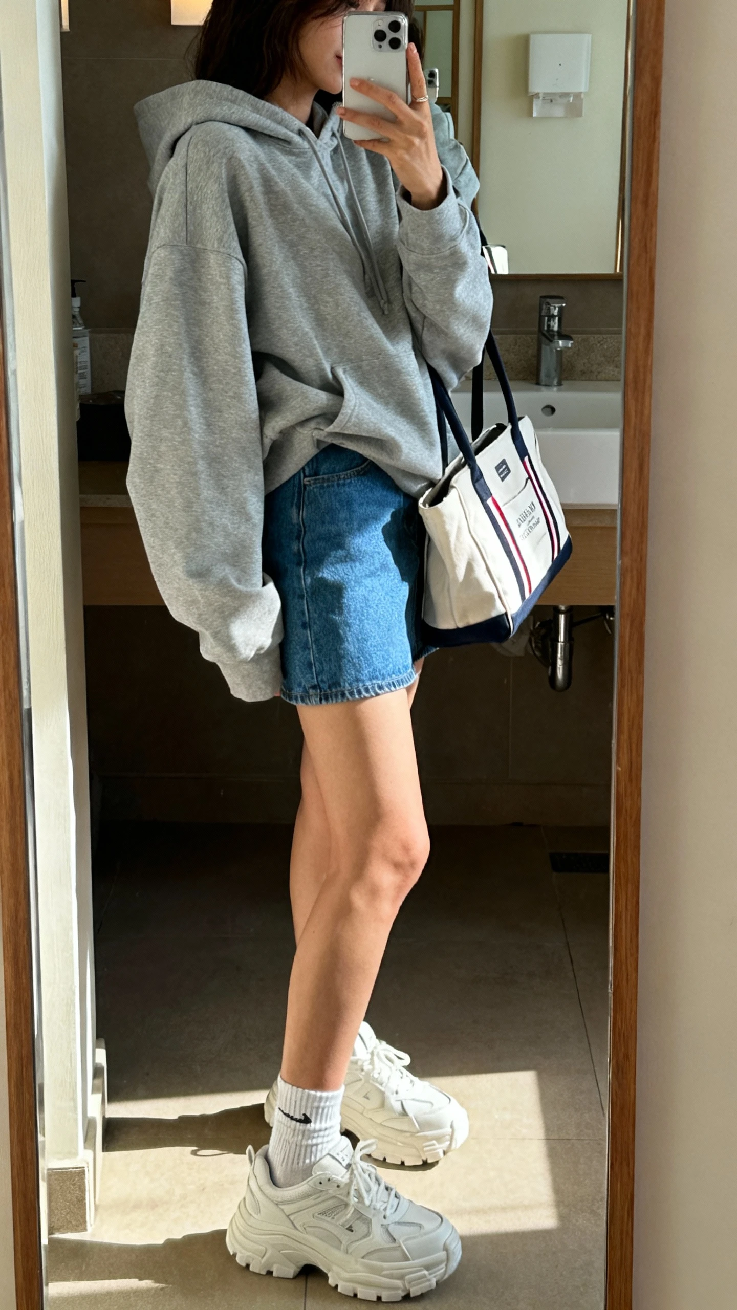 iPhone bathroom mirror selfie of a woman in an oversized gray hoodie, blue denim mini skirt, chunky white sneakers, and ankle socks with a sporty tote, face not visible, side angle, indoor lighting, iPhone photo quality.