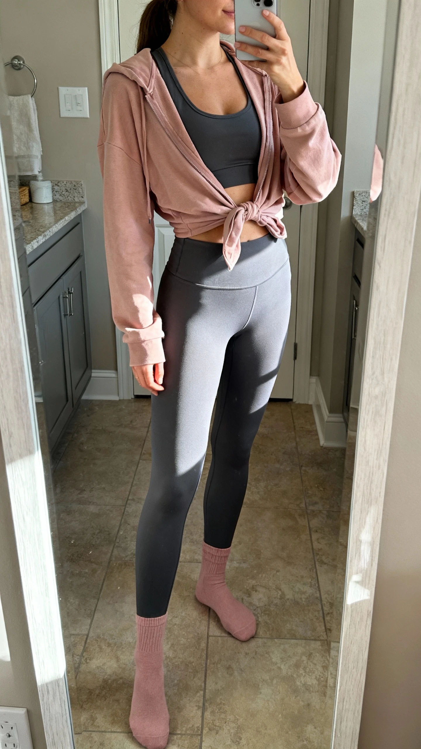 iPhone bathroom mirror selfie of a woman wearing charcoal gray leggings, a matching charcoal gray longline sports bra, and a soft blush pink lightweight hoodie tied at the waist with blush socks, face not visible, side angle, indoor morning light, iPhone photo quality.