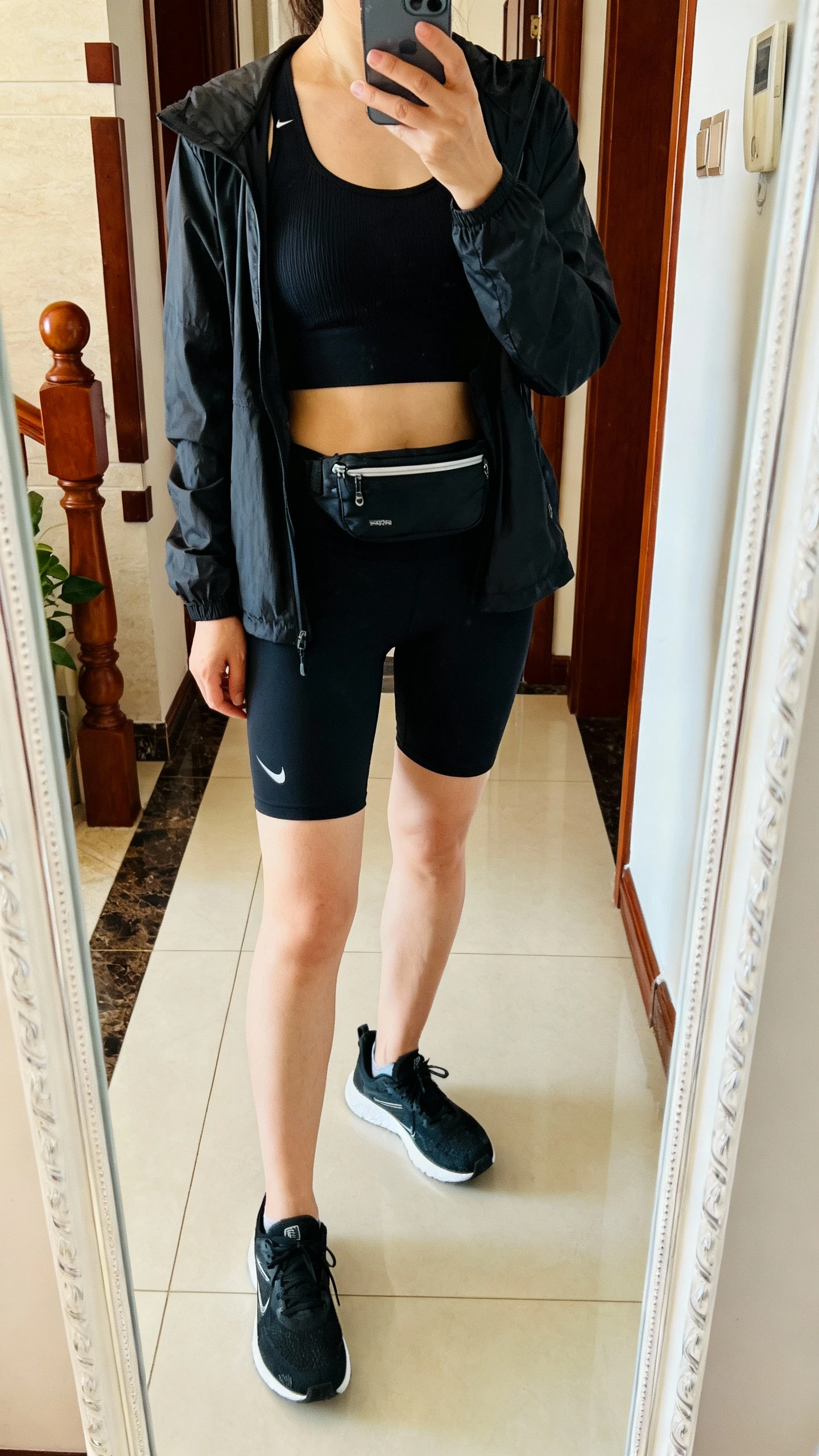 iPhone casual mirror selfie of a woman layering a lightweight black running jacket over a black breathable tank and black quick-dry shorts, with black running shoes and a small black waist pack, face not visible, entryway mirror, morning light, iPhone photo quality.
