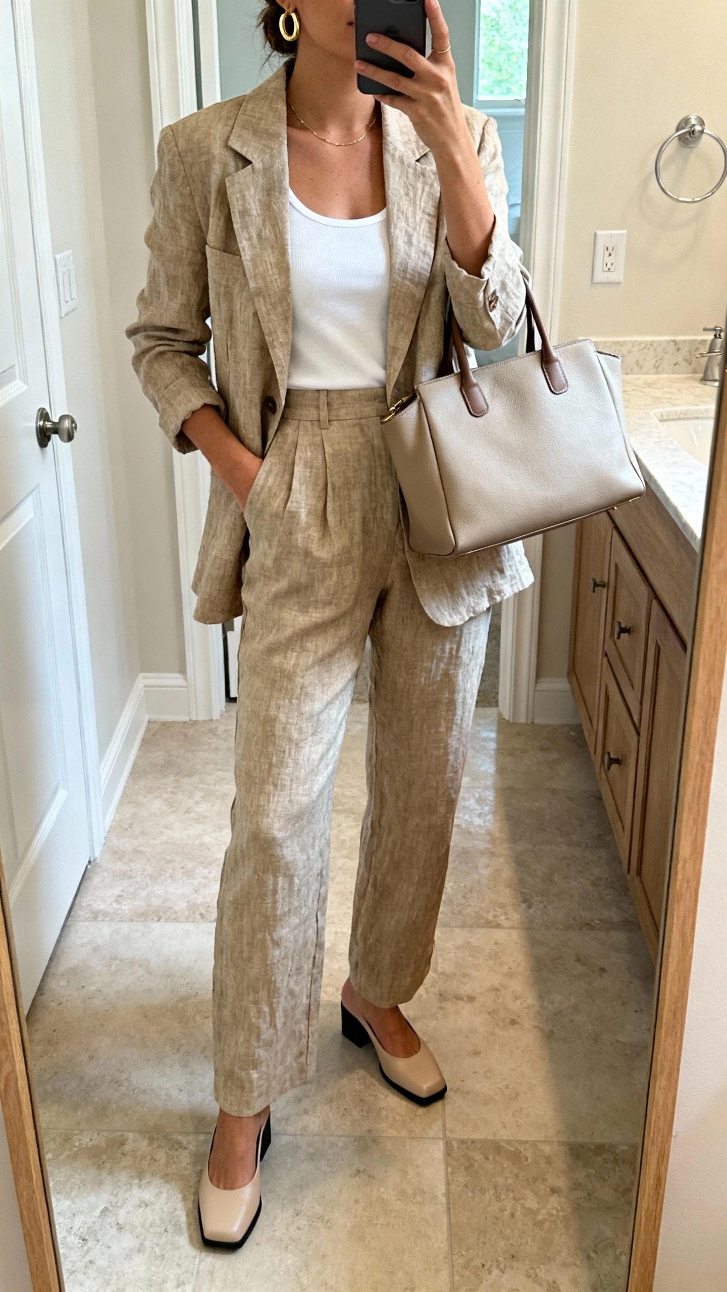 iPhone full-length mirror selfie of a woman in a linen blazer and matching high-waist trousers, white tank, low block heels, delicate gold hoops, and a structured tote, face not visible, bathroom mirror with soft indoor lighting, iPhone photo quality.