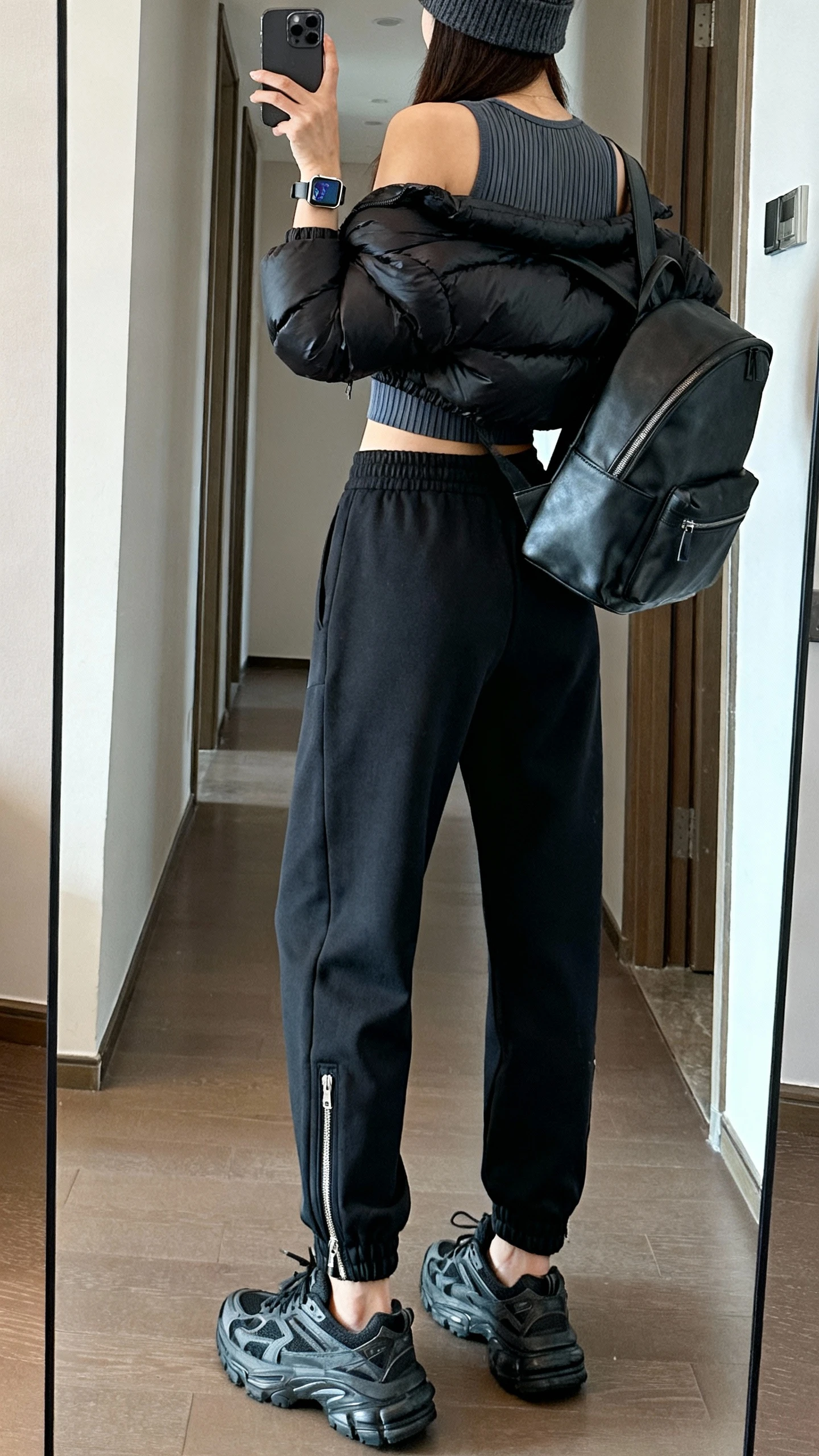 iPhone mirror selfie back view of a woman wearing high-rise track pants with zip hems, a fitted ribbed athletic tank, a black cropped puffer jacket, chunky dad sneakers, a beanie, a structured backpack, and a smartwatch; face not visible, hallway mirror, natural indoor light, iPhone photo quality.