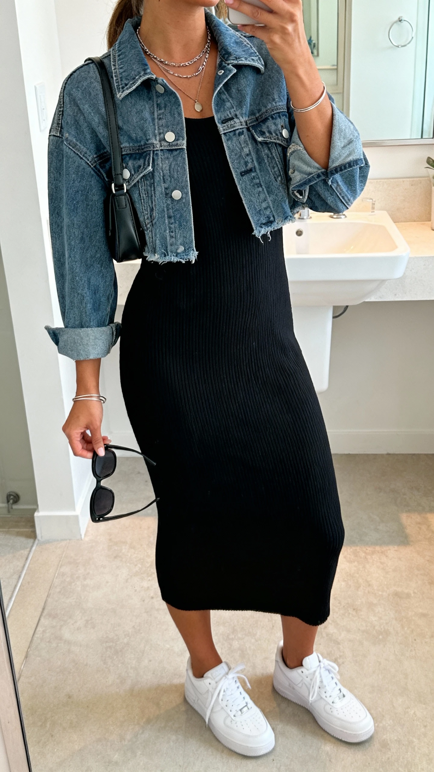 iPhone mirror selfie of a woman in a black ribbed knit midi dress with a cropped medium-wash denim jacket, clean white sneakers, small shoulder bag, layered necklaces, slim bangles, matte modern sunglasses in hand, face not visible, bathroom mirror, soft indoor lighting, iPhone photo quality.