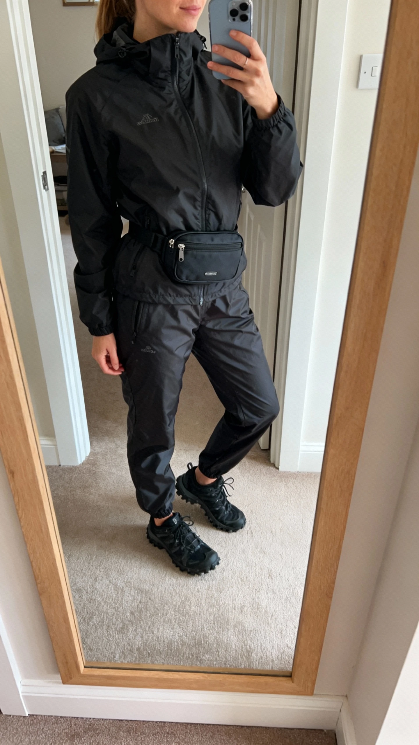 iPhone mirror selfie of a woman in a black windbreaker jacket and matching black windbreaker pants, face not visible, entryway mirror with natural daylight, black trail sneakers and a compact black waist pack, iPhone photo quality
