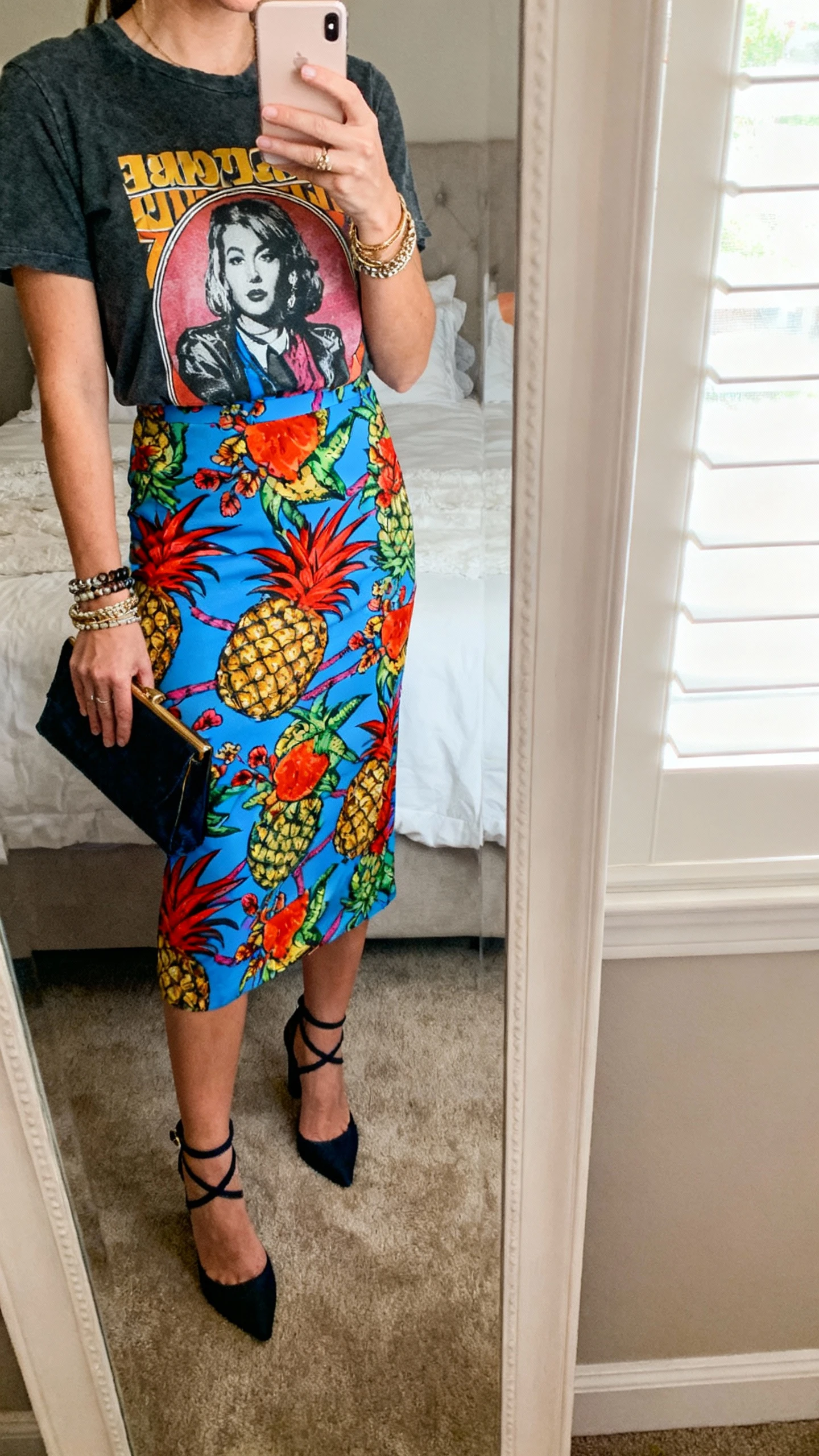 iPhone mirror selfie of a woman in a bold statement skirt (vibrant print) paired with a graphic tee, ankle-strap heels, layered bracelets, and a clutch, face not visible, cropped at neck, bedroom mirror with soft window light, iPhone photo quality.