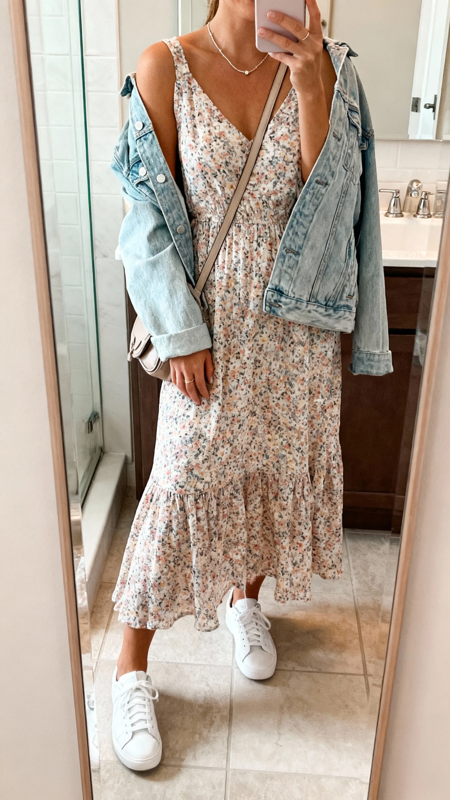 iPhone mirror selfie of a woman in a breezy floral midi dress with a light-wash denim jacket draped over shoulders, white sneakers, crossbody bag, and delicate necklace, face not visible, bathroom mirror, soft indoor lighting, iPhone photo quality.