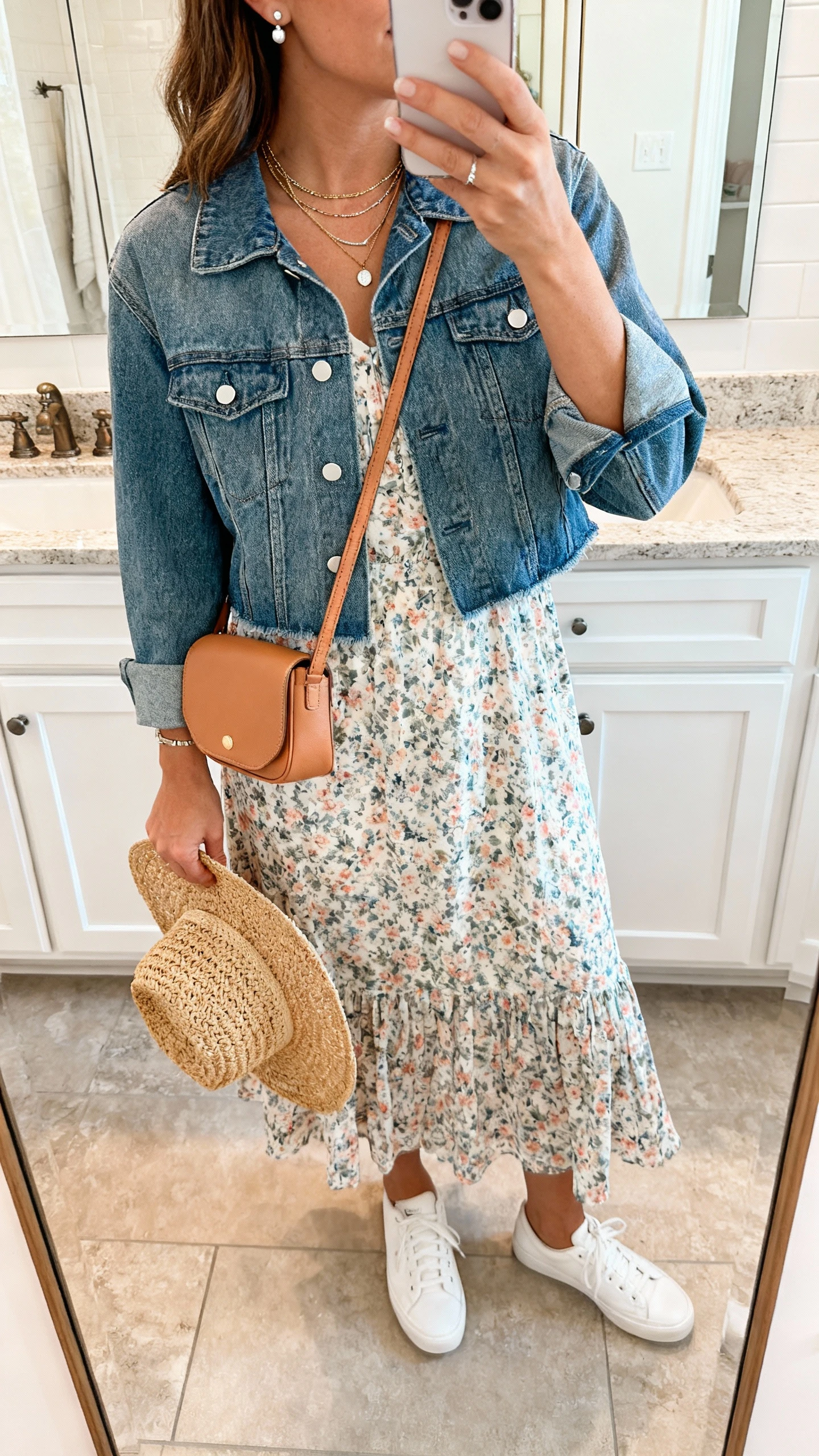 iPhone mirror selfie of a woman in a breezy floral midi dress with a slightly cropped medium-wash denim jacket, white sneakers, tan crossbody bag, layered necklaces, stud earrings, and a woven straw hat in hand; face not visible, bathroom mirror, natural indoor lighting, iPhone photo quality.