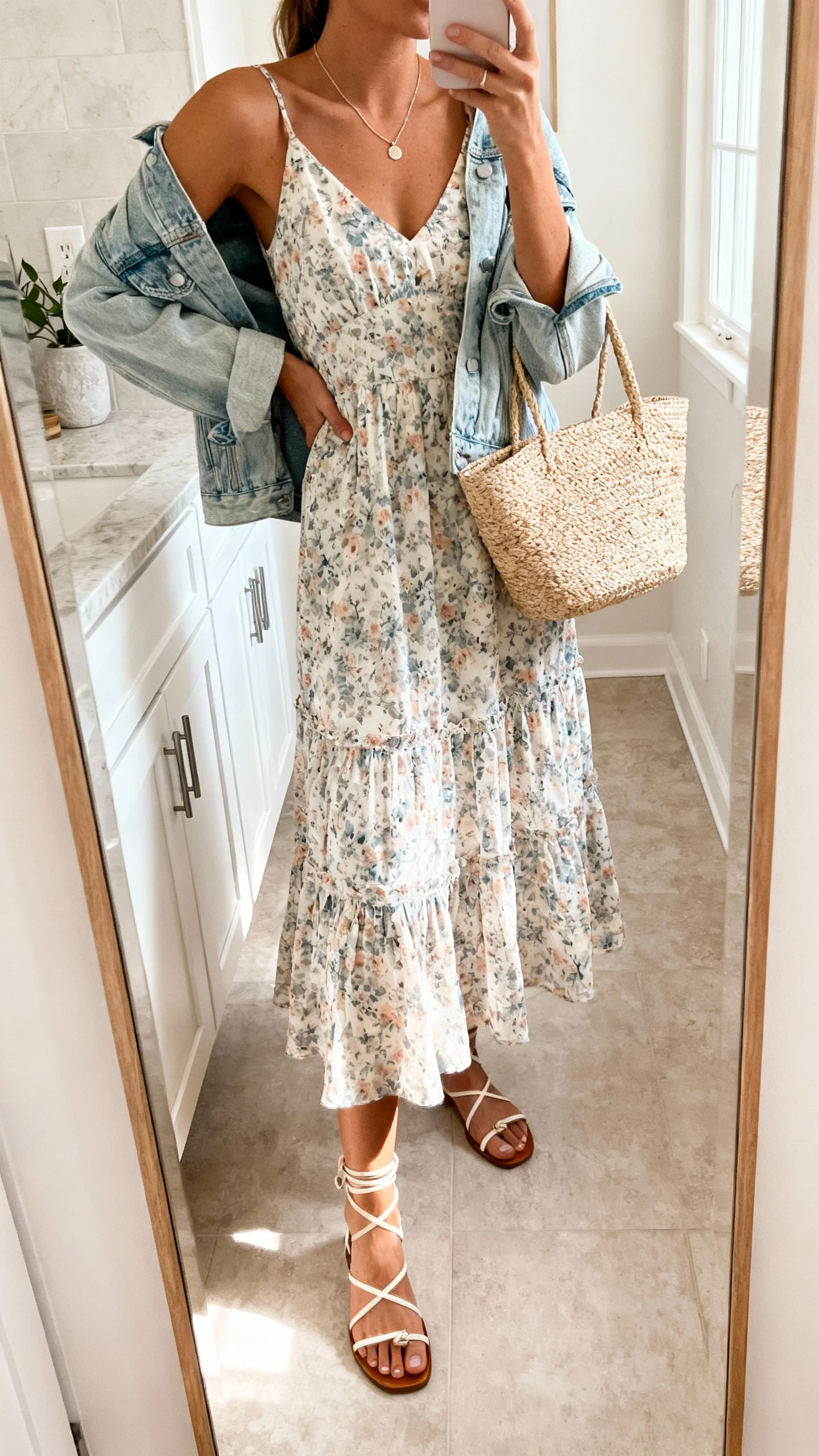 iPhone mirror selfie of a woman in a breezy floral midi dress with a light-wash denim jacket draped over shoulders, ankle-strap sandals, straw tote, delicate necklace, face not visible, bathroom full-length mirror, morning light, iPhone photo quality.