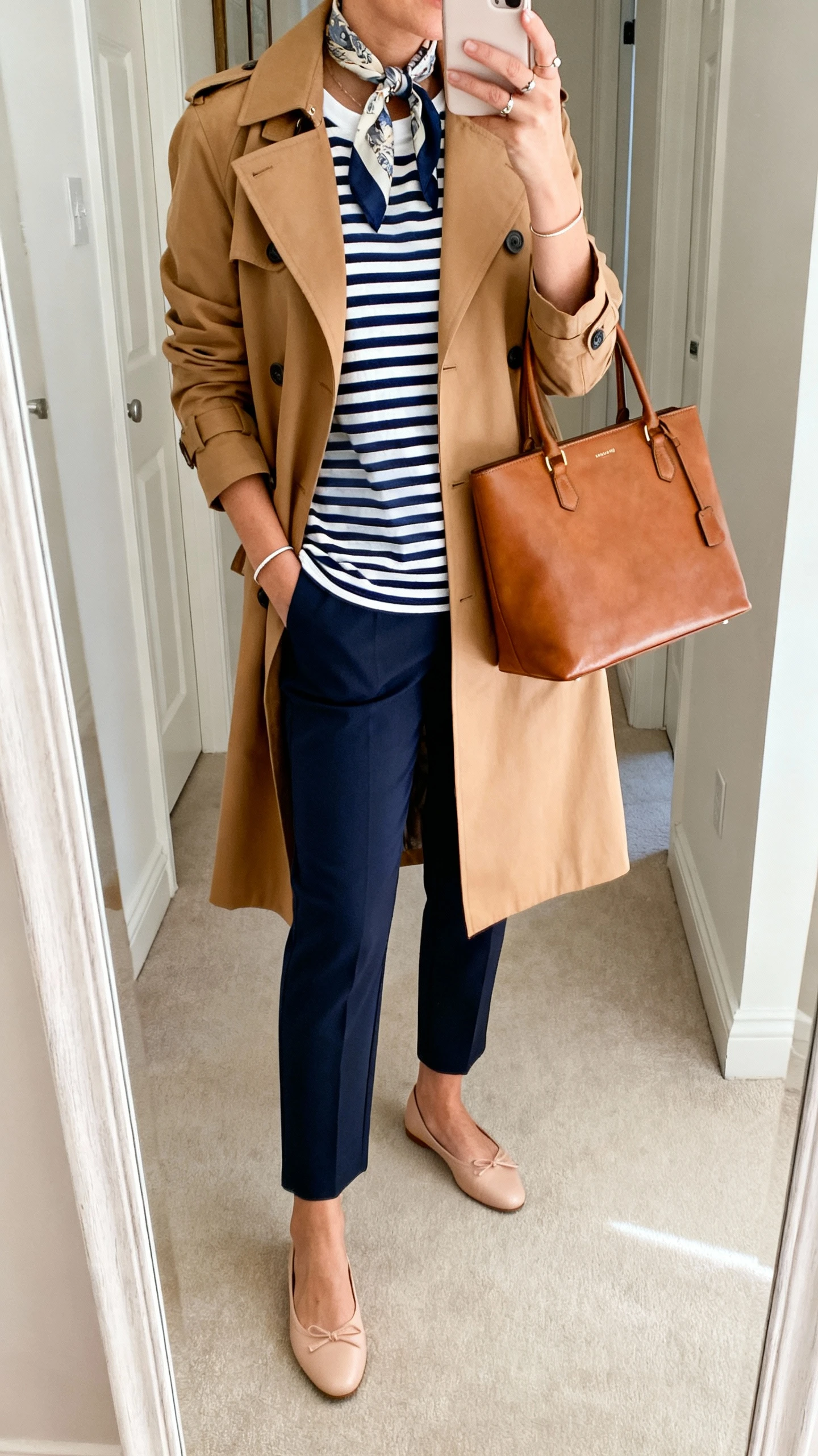 iPhone mirror selfie of a woman in a camel trench coat over a navy-and-white Breton striped tee, cropped straight trousers, ballet flats, structured leather tote, silk scarf tied at neck, simple studs, and a slim bracelet; face not visible, hallway mirror, soft indoor lighting, iPhone photo quality.