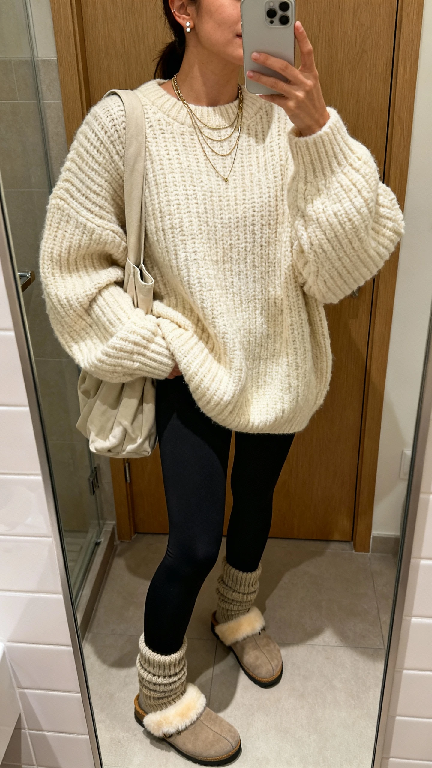 iPhone mirror selfie of a woman in a chunky oversized knit sweater in cream, matte black leggings, shearling-lined clogs, thick socks peeking out, a slouchy shoulder bag, delicate layered necklaces and stud earrings, face not visible, bathroom mirror, warm indoor lighting, iPhone photo quality.