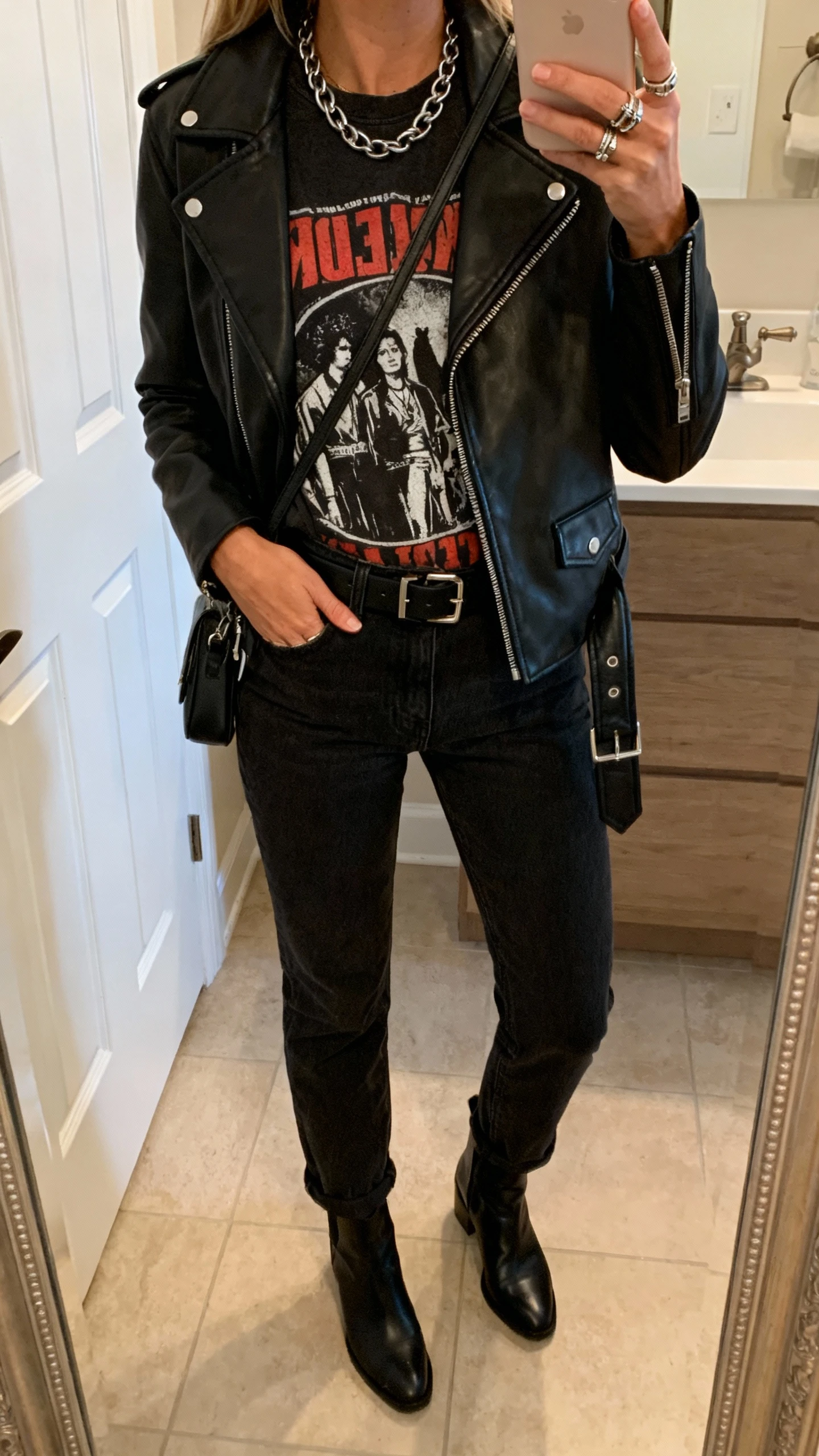 iPhone mirror selfie of a woman in a classic black leather moto jacket, graphic band tee, black mom jeans, ankle boots, a chain necklace, stacked rings, black belt, and a small crossbody, face not visible, bathroom mirror, indoor warm light, iPhone photo quality.