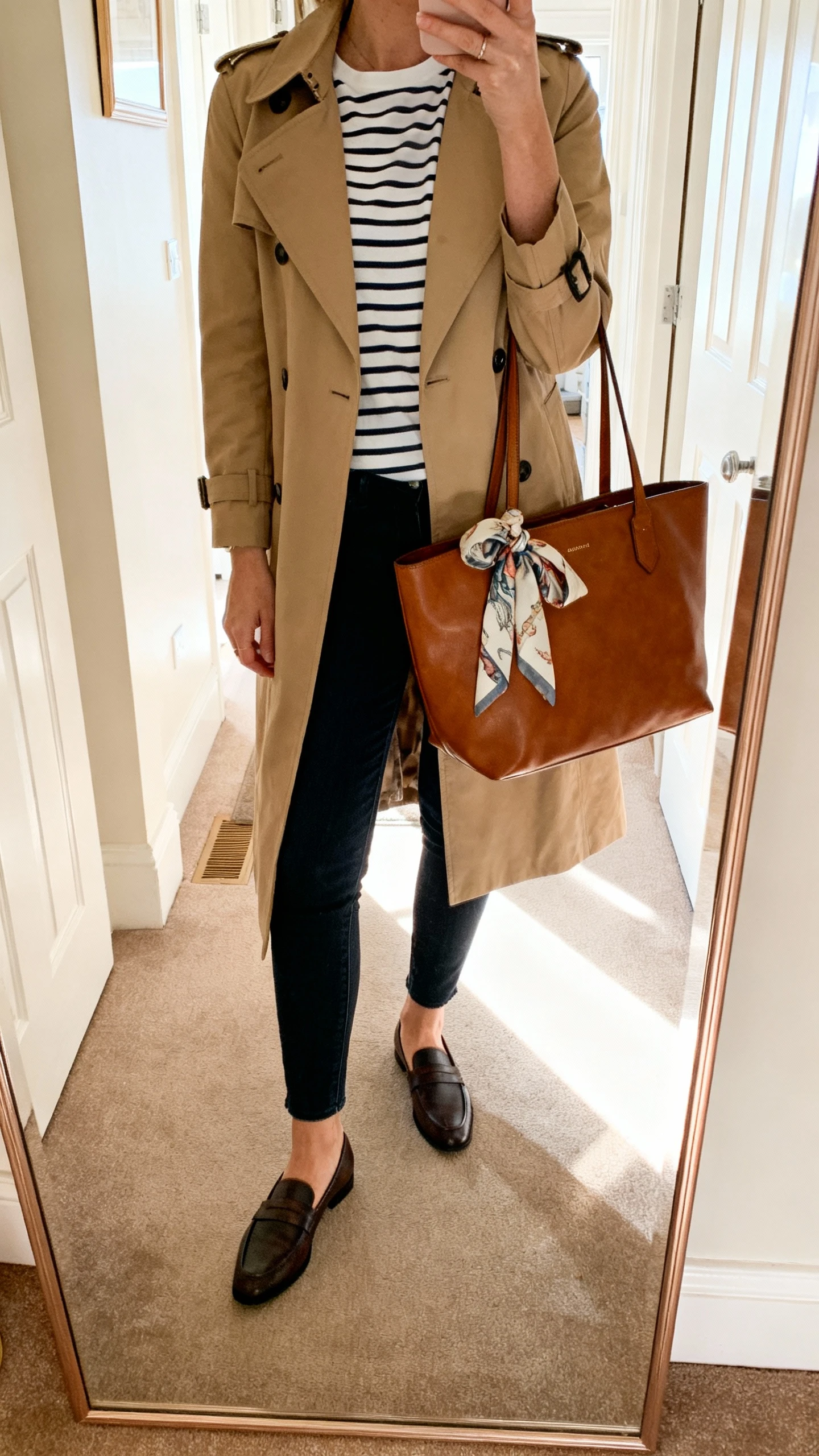 iPhone mirror selfie of a woman in a classic trench coat over a Breton striped tee, slim dark jeans, loafers, leather tote, silk scarf tied to handle, face not visible, cropped at neck, entryway full-length mirror, morning light, iPhone photo quality.