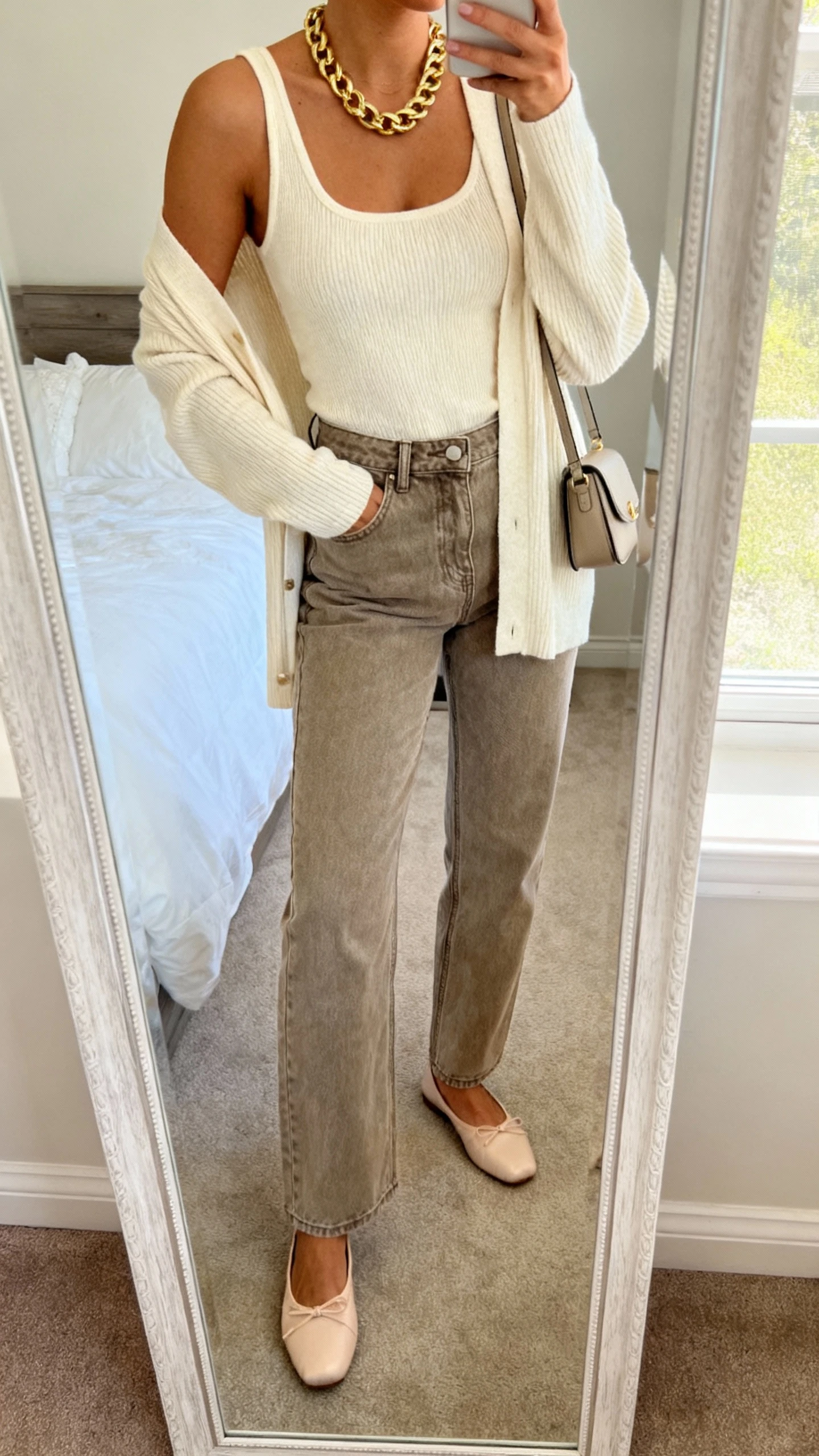 iPhone mirror selfie of a woman in a cream cardigan set (fine-knit tank and matching cardigan), high-waist stone jeans, chunky gold chain, ballet flats, small shoulder bag, face not visible, cropped at neck, bedroom mirror, natural window light, iPhone photo quality.