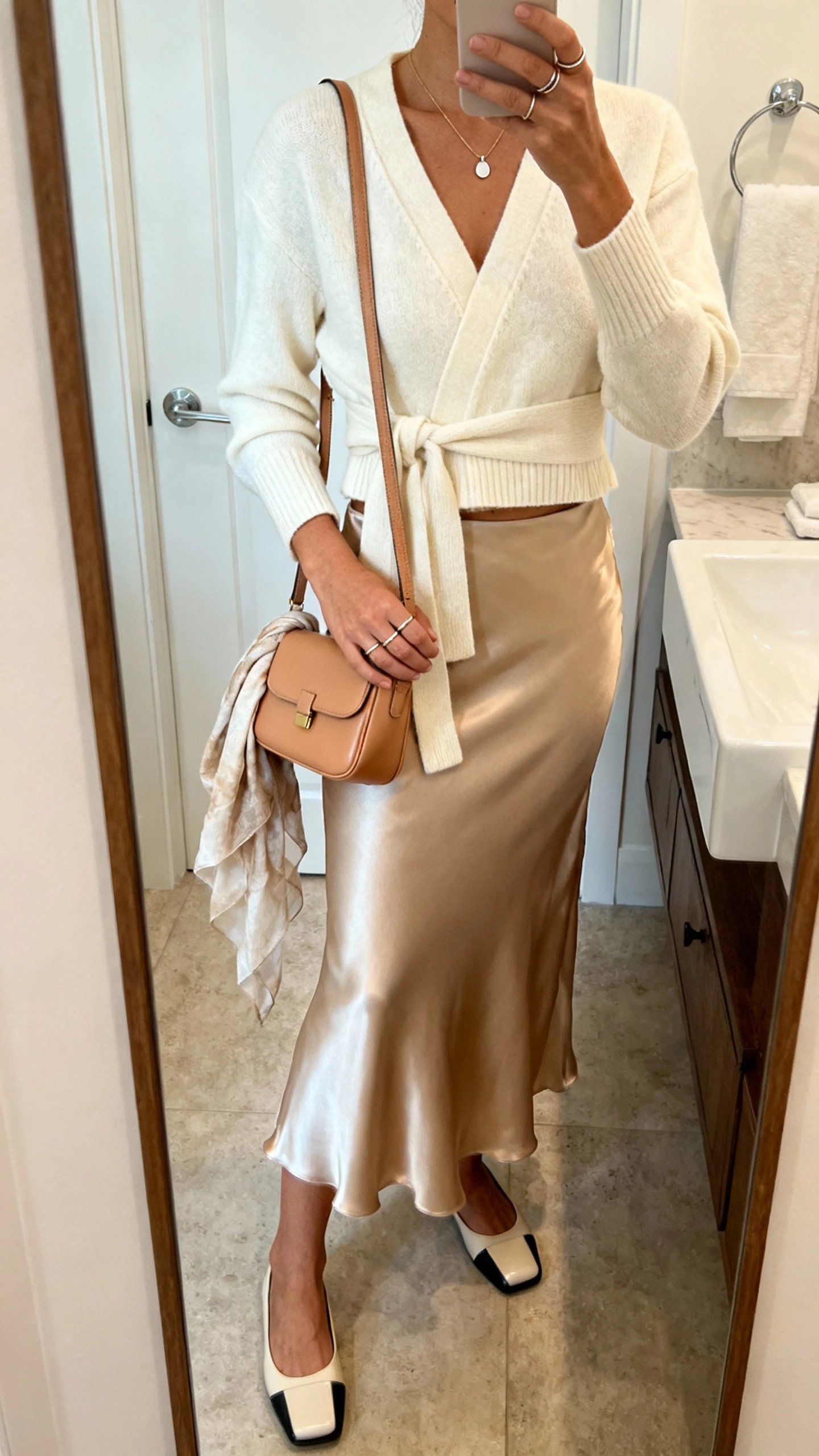 iPhone mirror selfie of a woman in a cream cropped wrap cardigan, champagne satin slip skirt, square-toe ballet flats, fine pendant necklace, thin stacking rings, mini tan shoulder bag, and a lightweight scarf draped over the bag, face not visible, bathroom mirror with soft indoor light, iPhone photo quality.