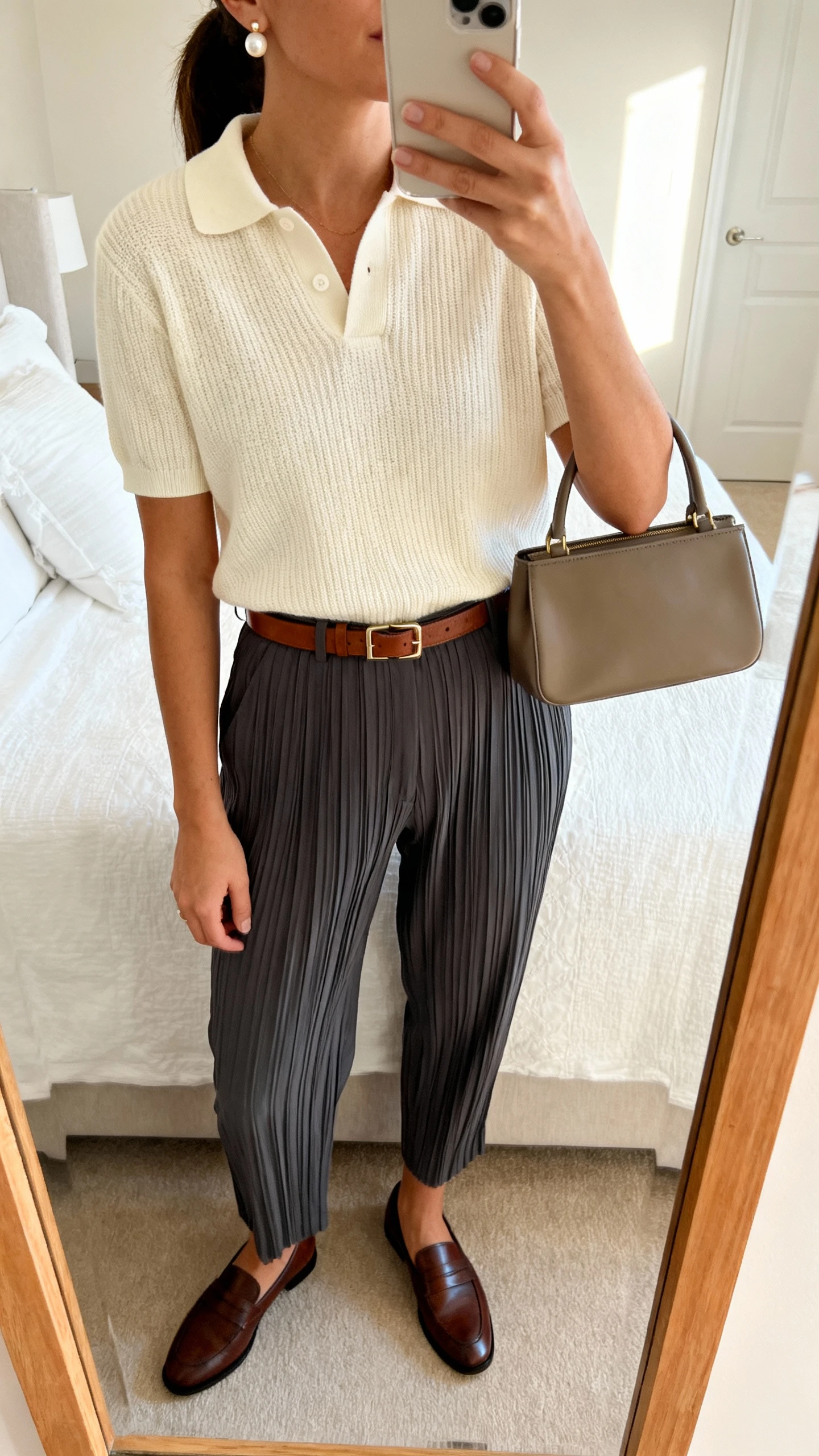 iPhone mirror selfie of a woman in a cream knit polo tucked into charcoal pleated trousers, leather loafers, slim leather belt, structured top-handle bag, pearl studs, face not visible, cropped at neck, bedroom mirror, soft afternoon light, iPhone photo quality.